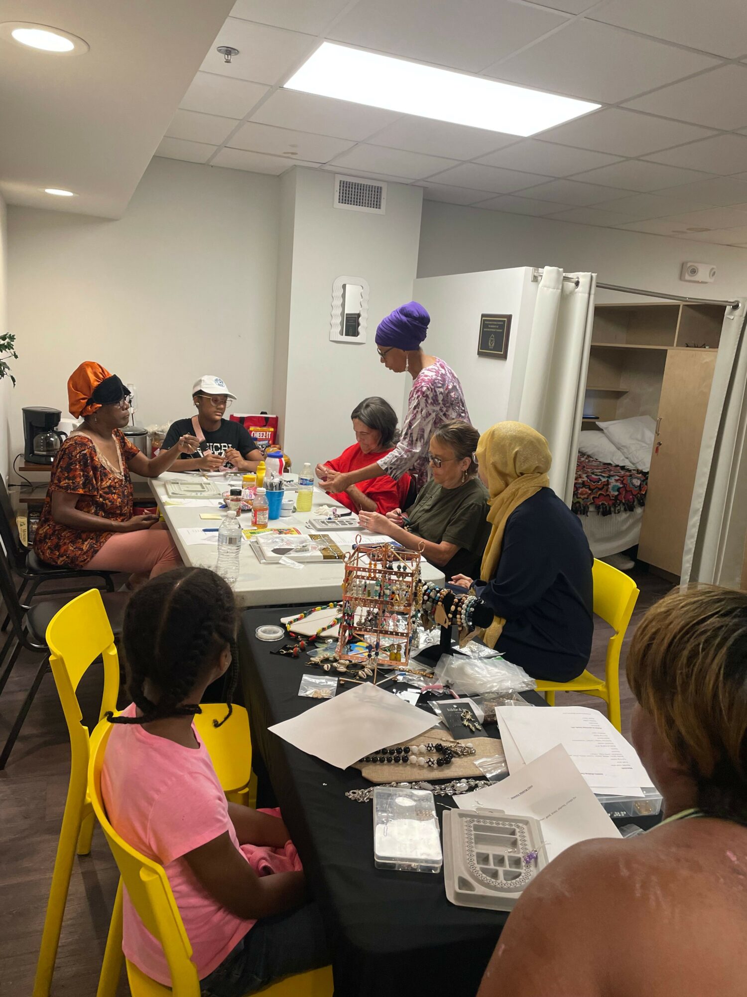 Group of women and children sitting around a table with food and drinks in a room with white walls and ceiling lights.