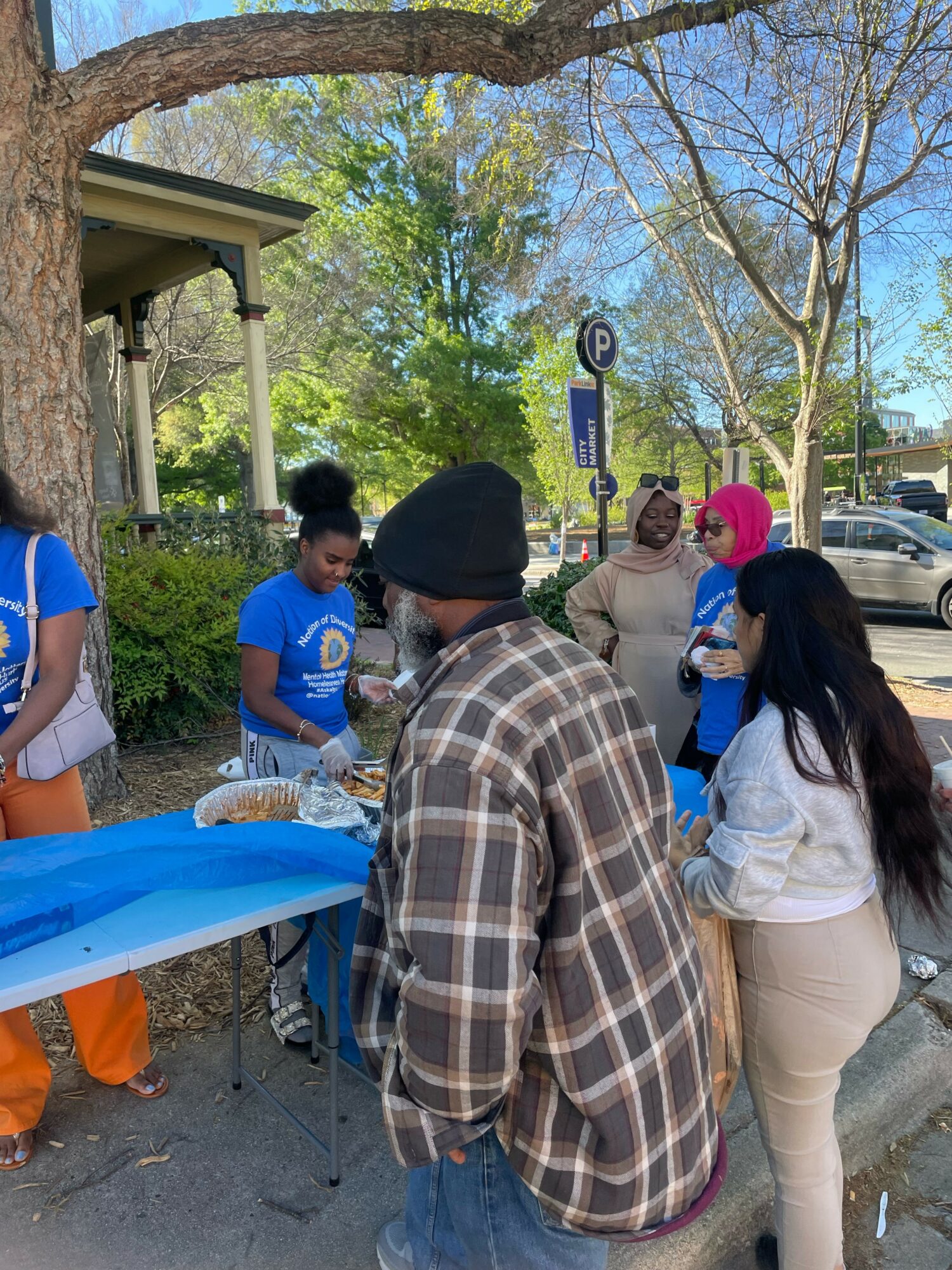 Group of people gathered outdoors near a table with food, trees, and parked cars in background.