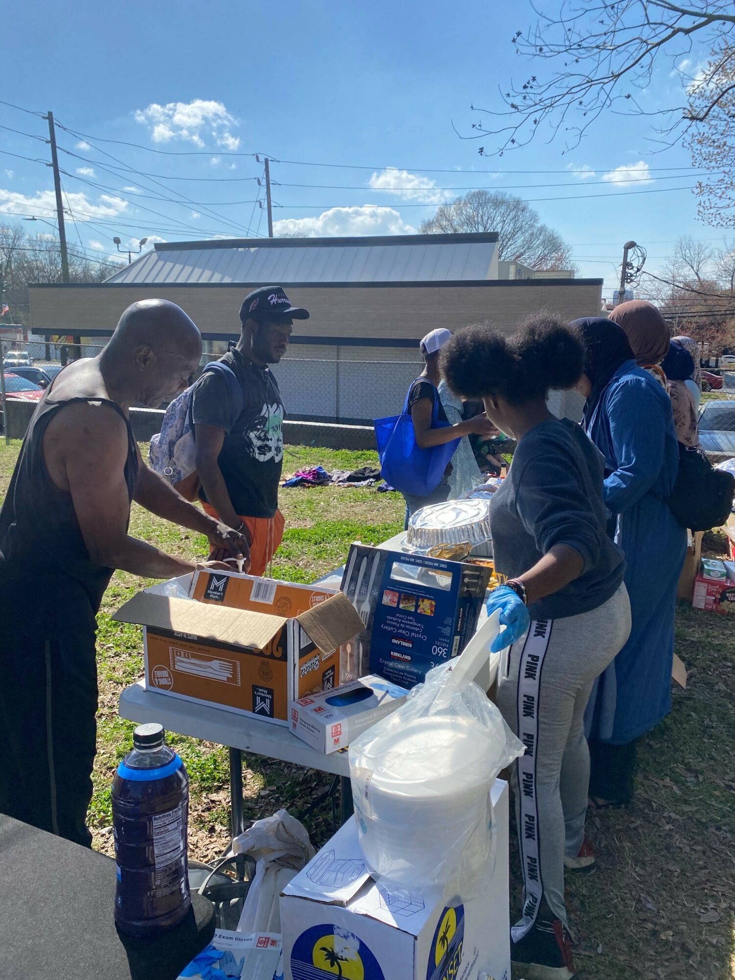 Group of people gathered around a table outdoors, some with backpacks, in a sunny area with a building and trees in background.