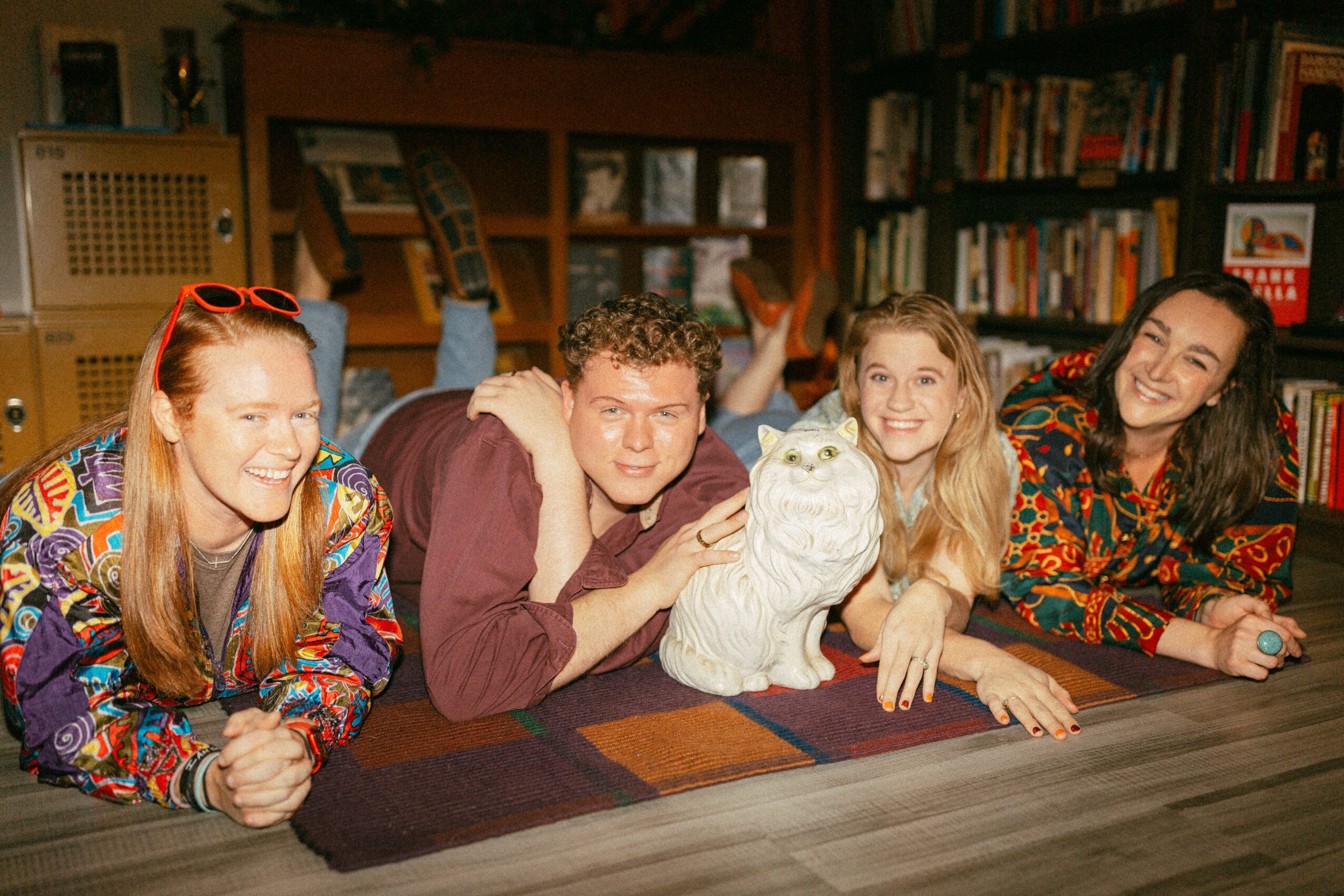 Four people lying on a table in a room with bookshelves, smiling and looking at the camera, with a white cat.