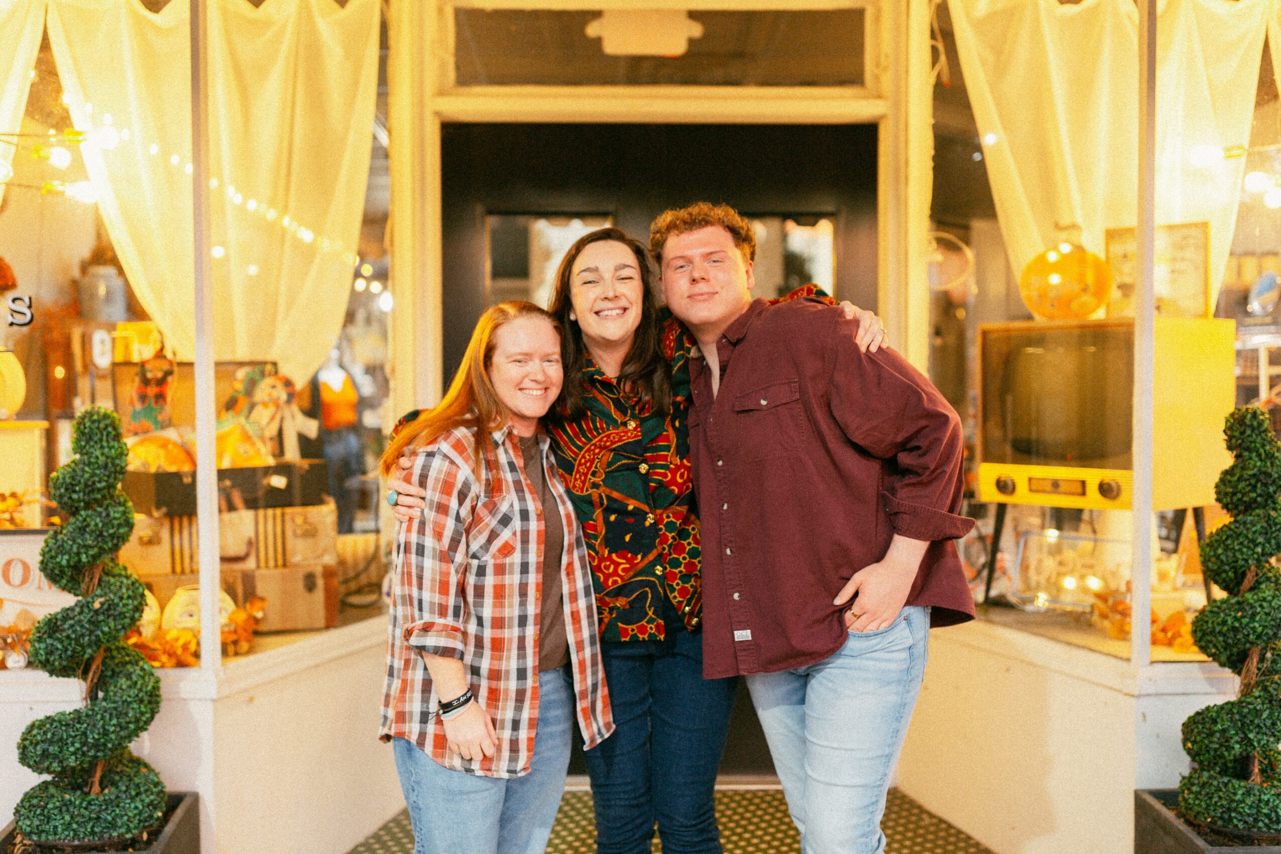 Three people smiling and standing close together indoors with yellow curtains and decorative plants behind them.