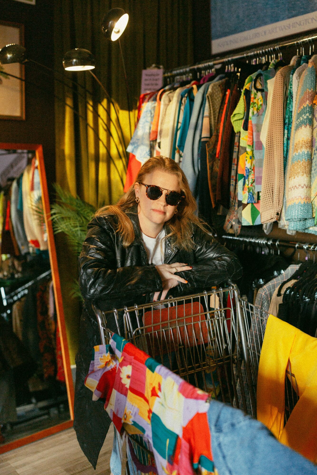 Woman with sunglasses and leather jacket shopping in a clothing store with colorful garments and a shopping cart.
