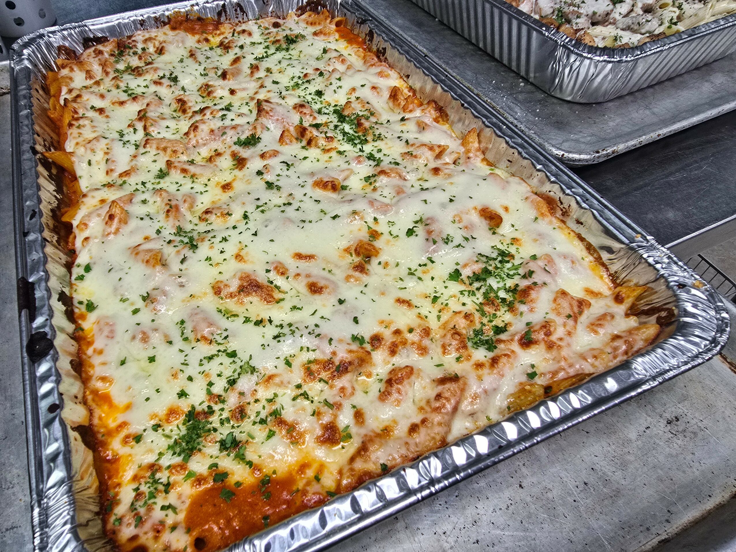 Large rectangular baked cheese pizza with herbs in a foil tray, with another tray of food partially visible behind.