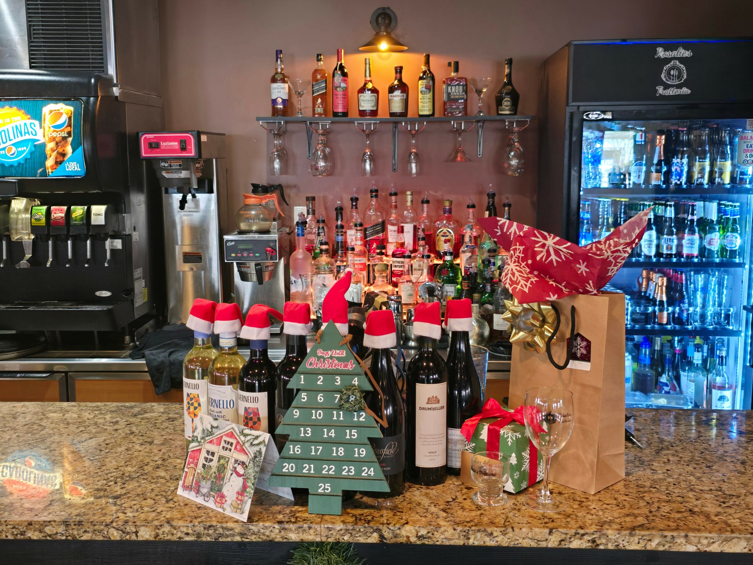 Bar counter with Christmas decorations, gift bags, bottles, and a small Christmas tree, with beverage machines and a refrigerator in background.