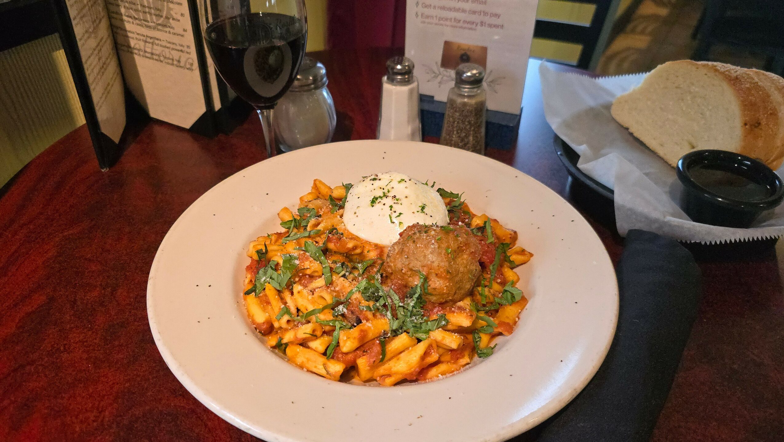 Plate with pasta, topped with a meatball, garnished with herbs and a dollop of sauce, on a wooden table.