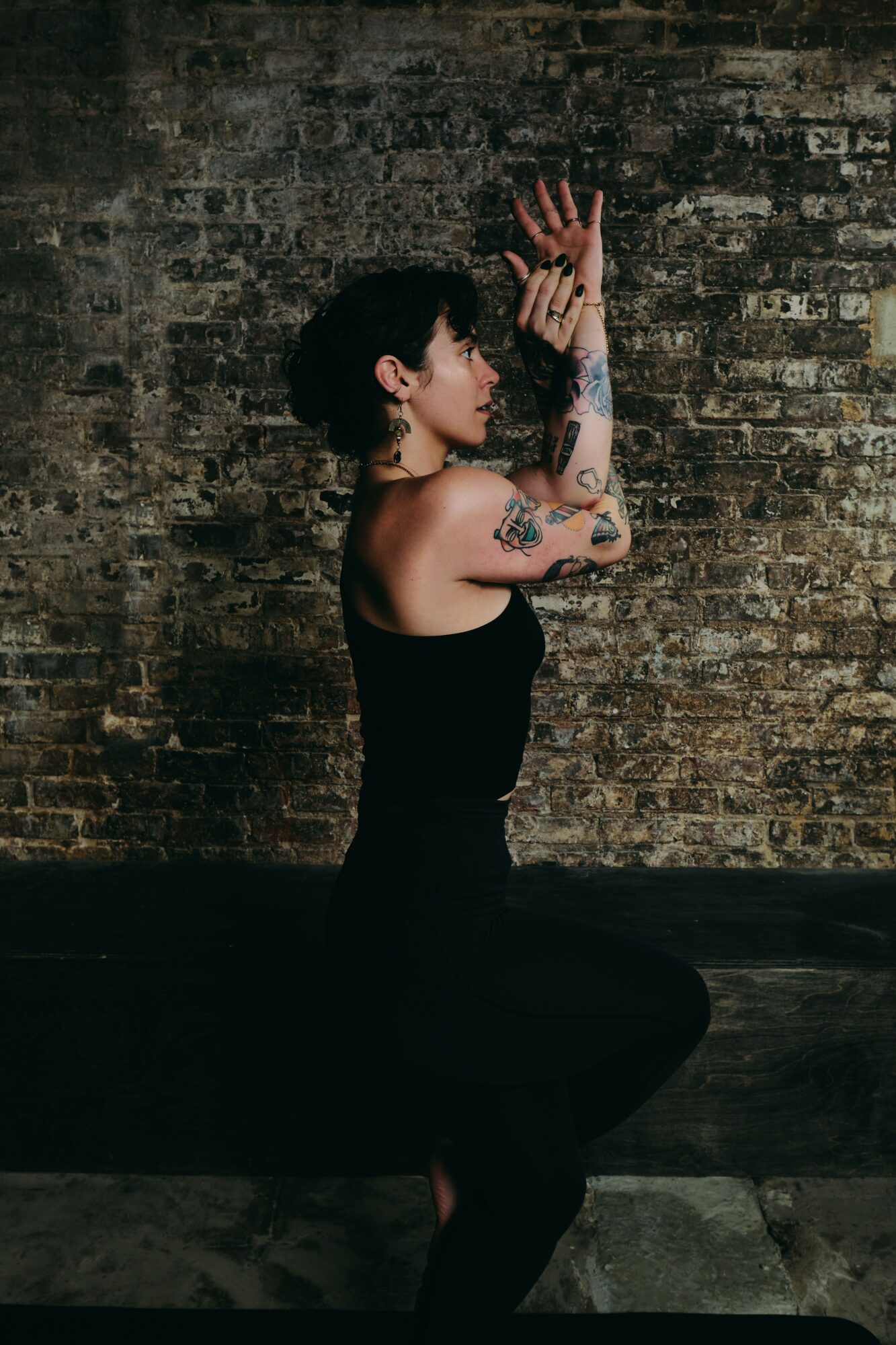 Woman with tattoos in black dress poses with hands raised against a brick wall.