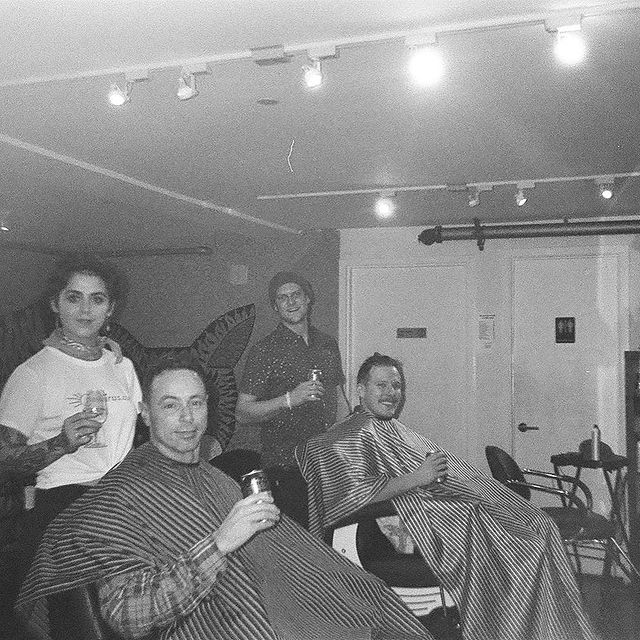 Four people in a room, two seated with capes, two standing, holding drinks, smiling, with a plain wall and ceiling lights.
