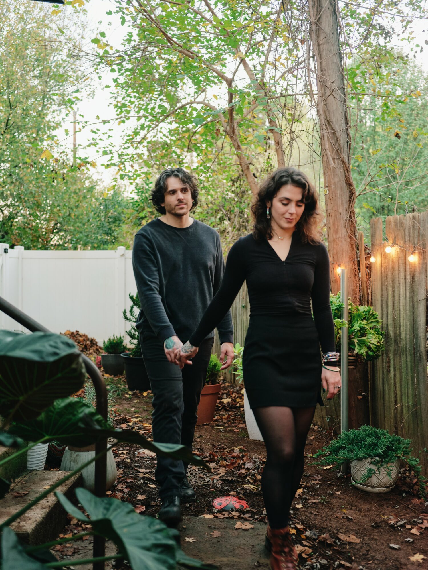 A woman in a black dress and patterned tights walks in a garden with a man in a dark sweater behind her.