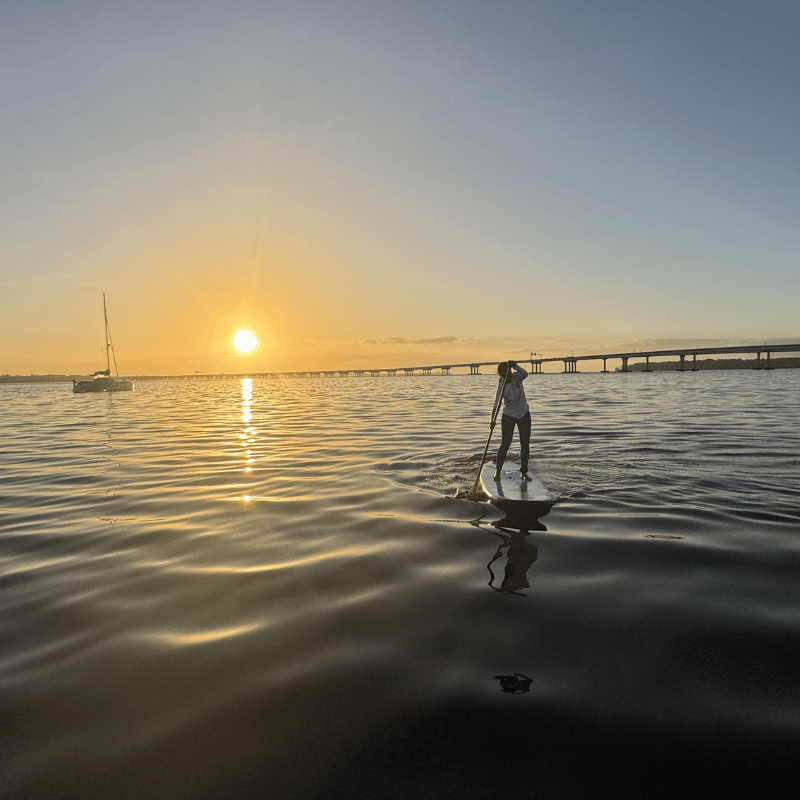 Person paddleboarding on water during sunset with a bridge in the background.