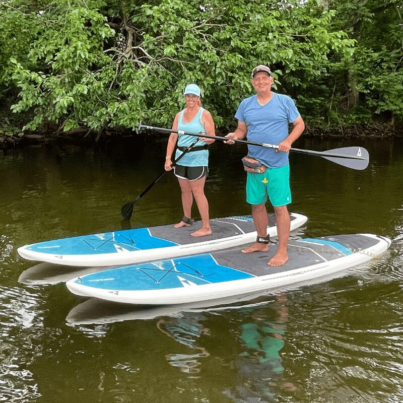 Two people stand on paddleboards in water, holding paddles, with green trees in the background.
