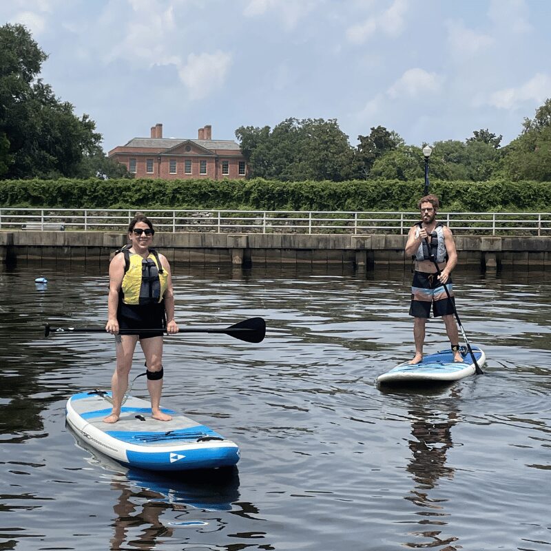 Two people stand on paddleboards in water, wearing life jackets and casual clothing, with trees and buildings in background.