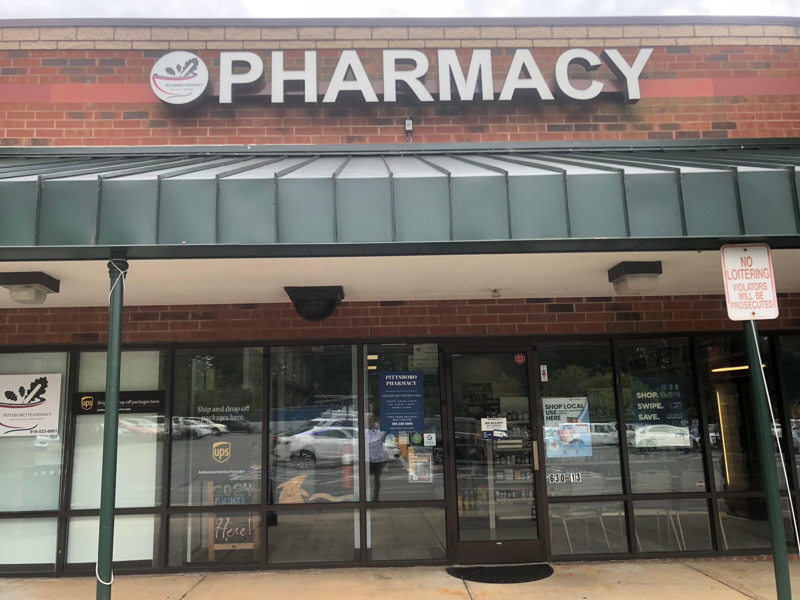 Pharmacy storefront with glass doors and windows, brick building, and a green awning above the entrance.