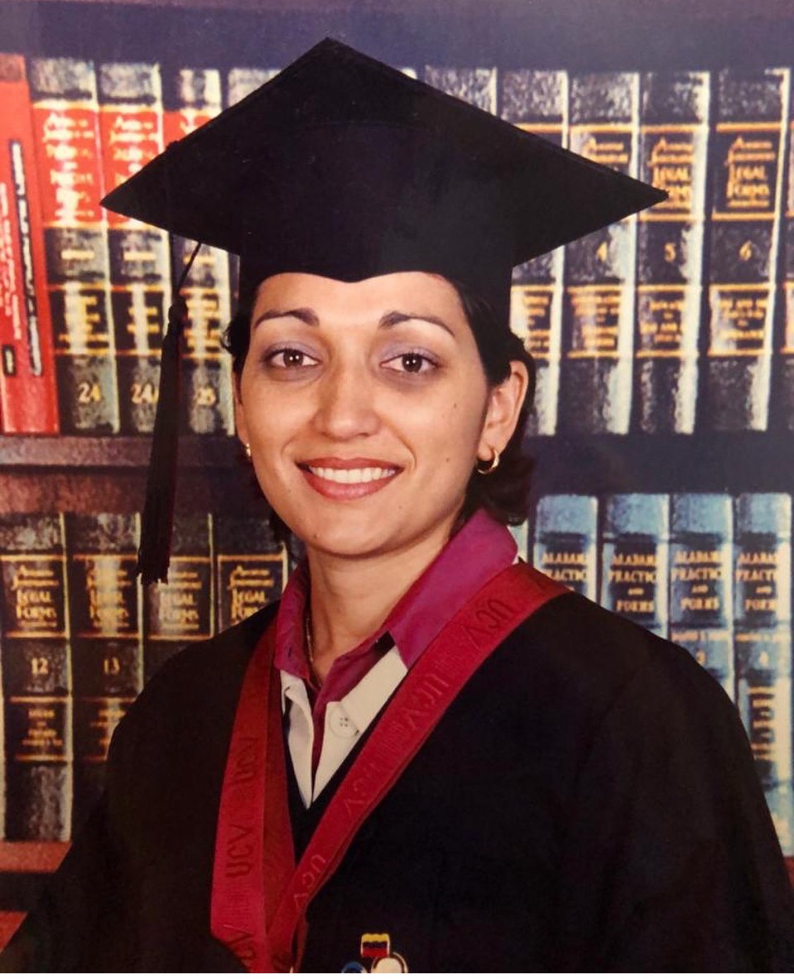 Woman in graduation cap and gown smiling, standing in front of a bookshelf filled with books.