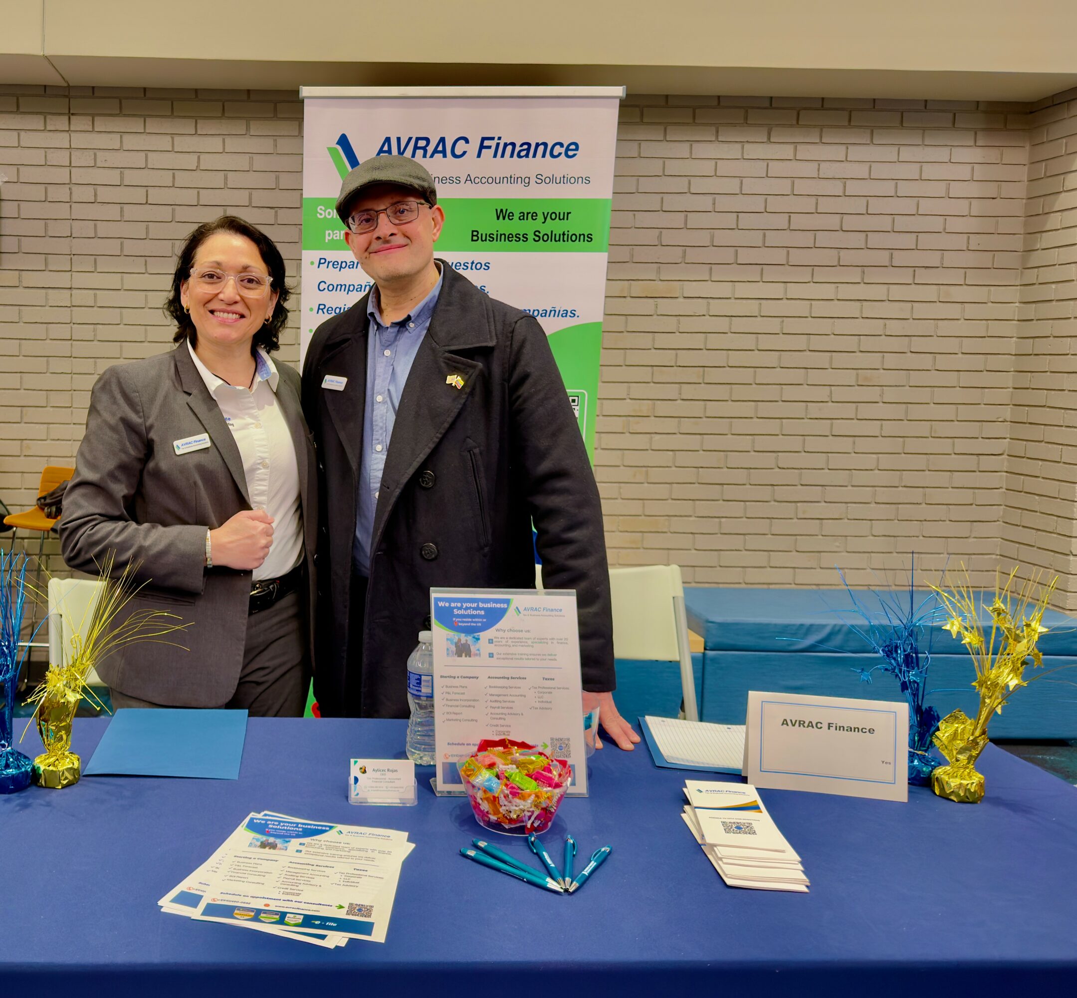 Two people standing behind a table at a booth, smiling, with a banner and promotional materials, in an indoor setting.
