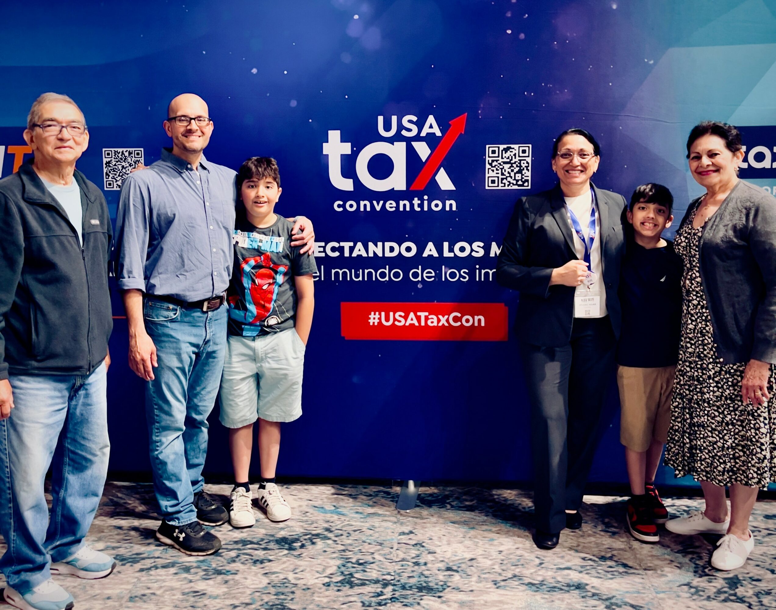 Group of seven people standing in front of a USA Tax convention backdrop, smiling, indoors.