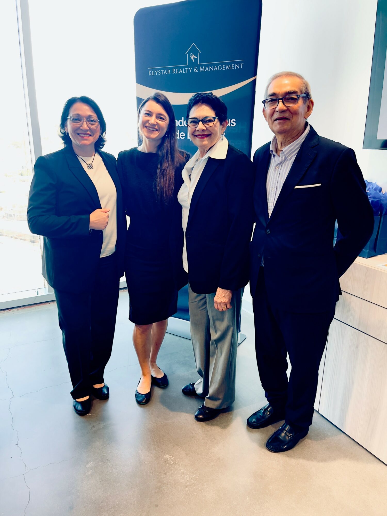 Four professionally dressed people standing together indoors, smiling, with a blue banner in the background.