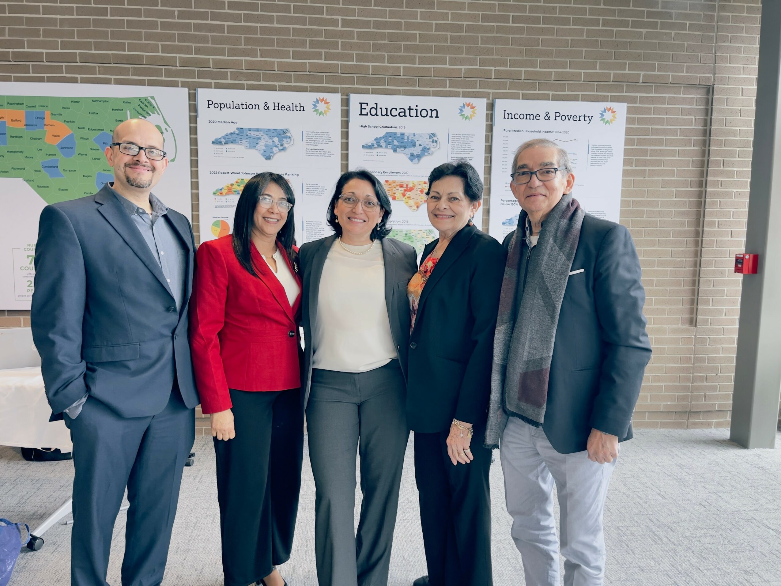 Five people standing in front of educational posters, smiling, indoors, casual and business attire, group photo.