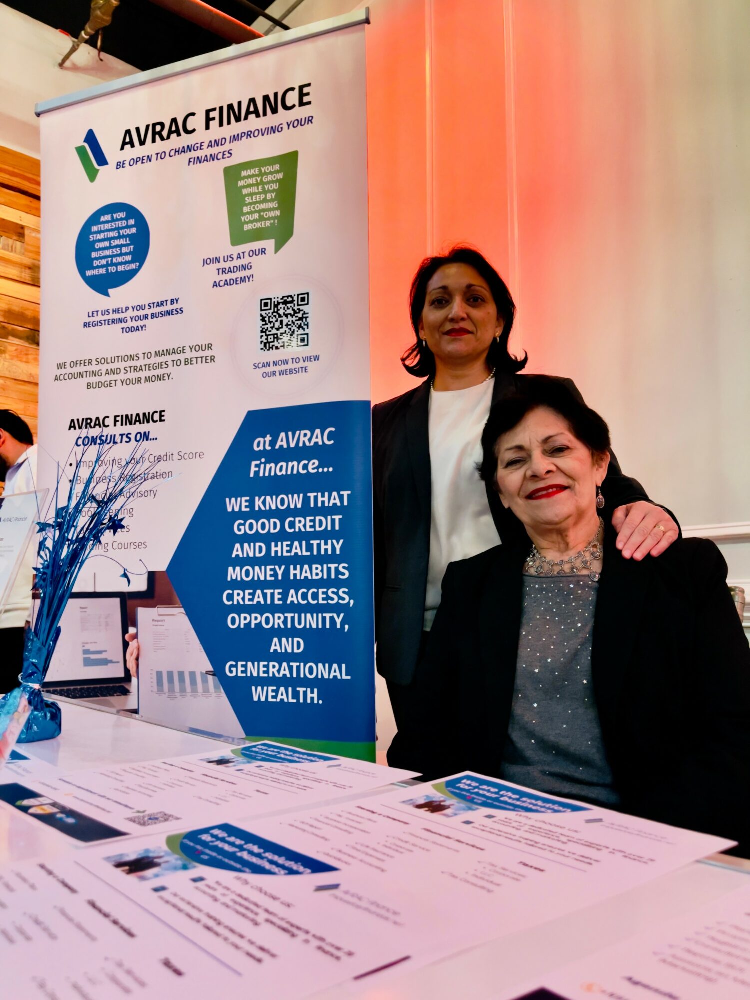 Two women standing behind a table with promotional materials at an event, with a large AVAC FINANCE banner in the background.