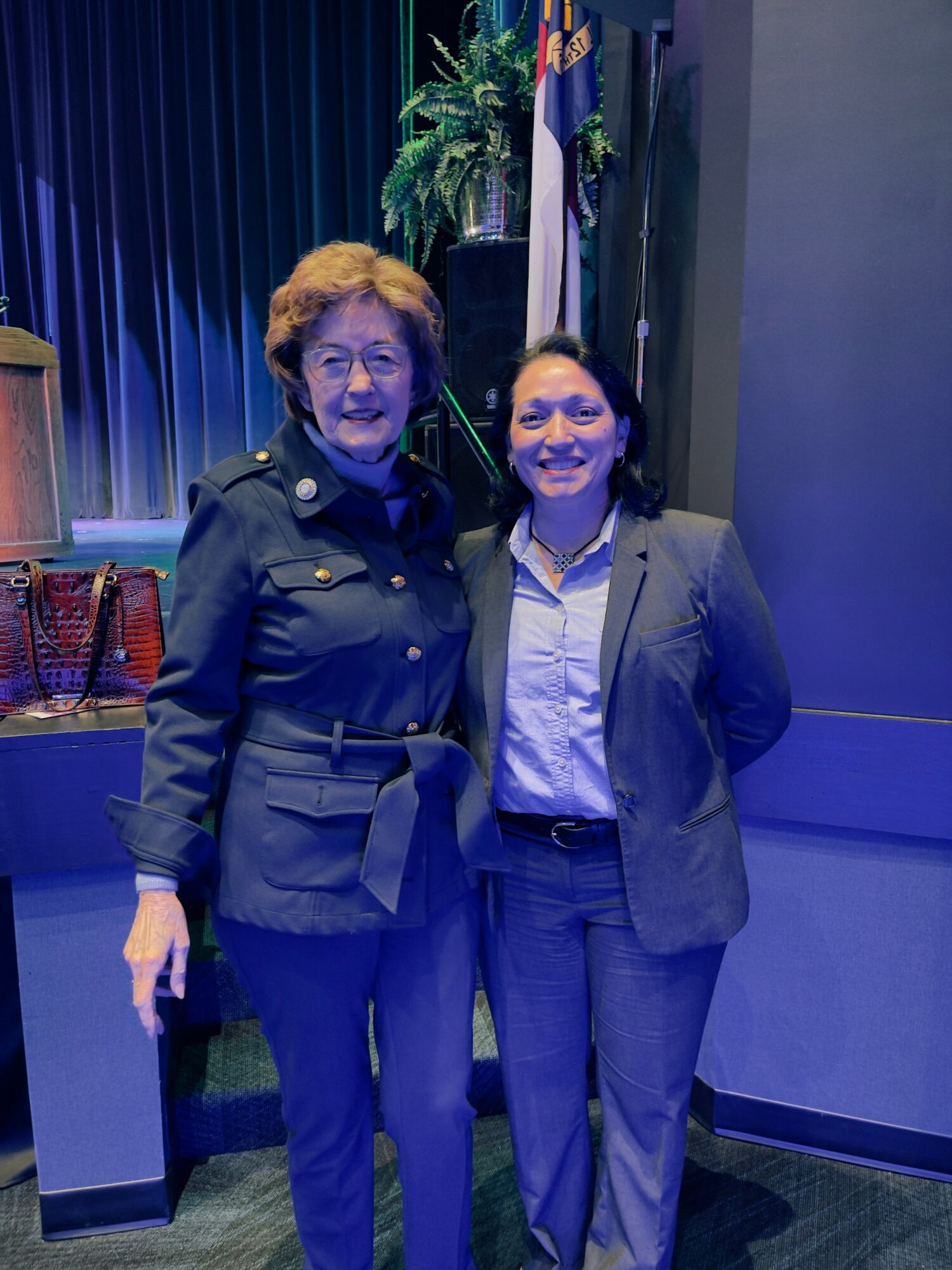 Two women standing together, smiling, in a room with blue curtains and plants, one in a dark jacket and the other in a suit.