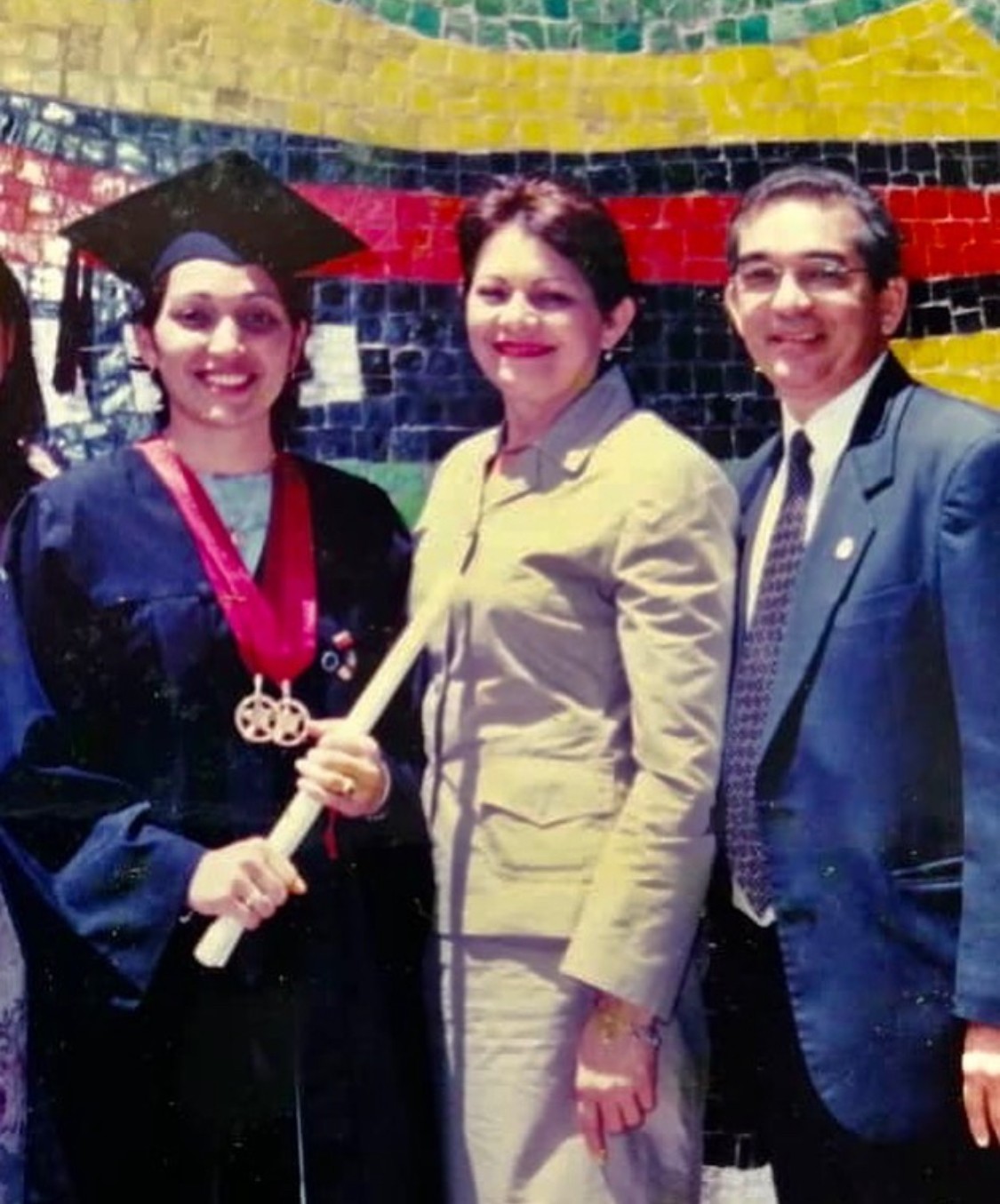Three people standing together, one in graduation attire holding a diploma, in front of a colorful mosaic wall.