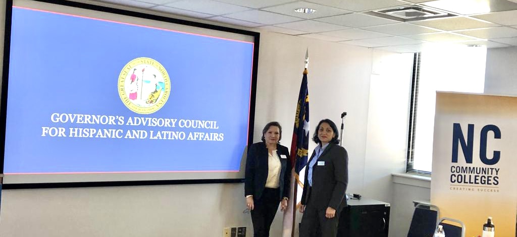 Two women standing in front of a large screen displaying the Governor's Advisory Council for Hispanic and Latino Affairs logo, in a conference room.
