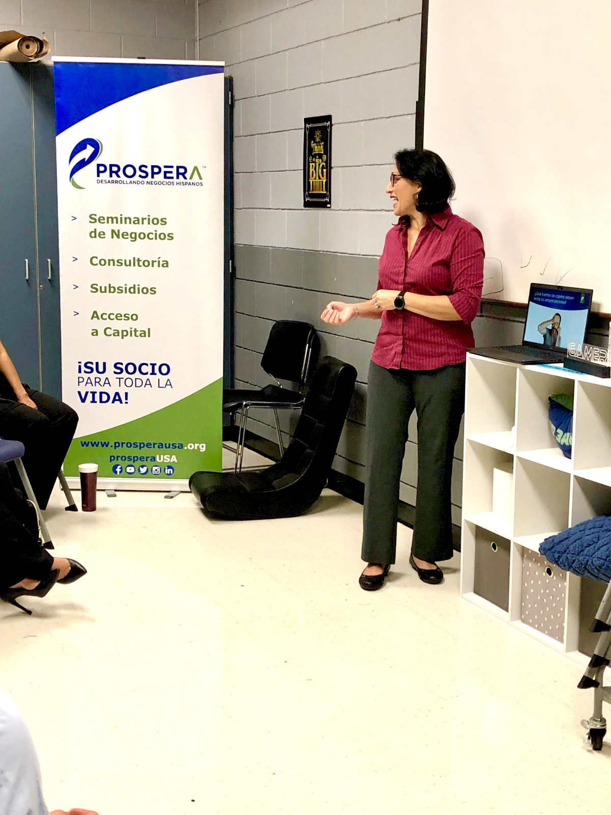 Woman in pink blazer and black pants standing near a white shelf, with a blue and white banner in the background, in an indoor setting.