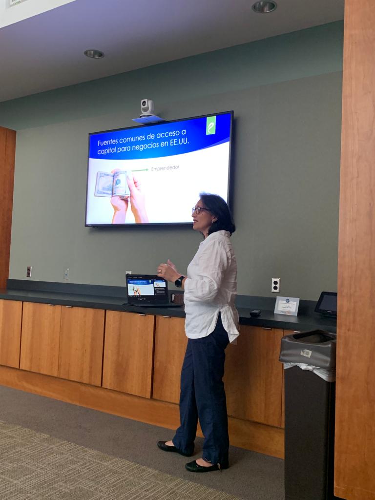 Woman standing in front of a large screen displaying a presentation, with a laptop on the counter beside her.