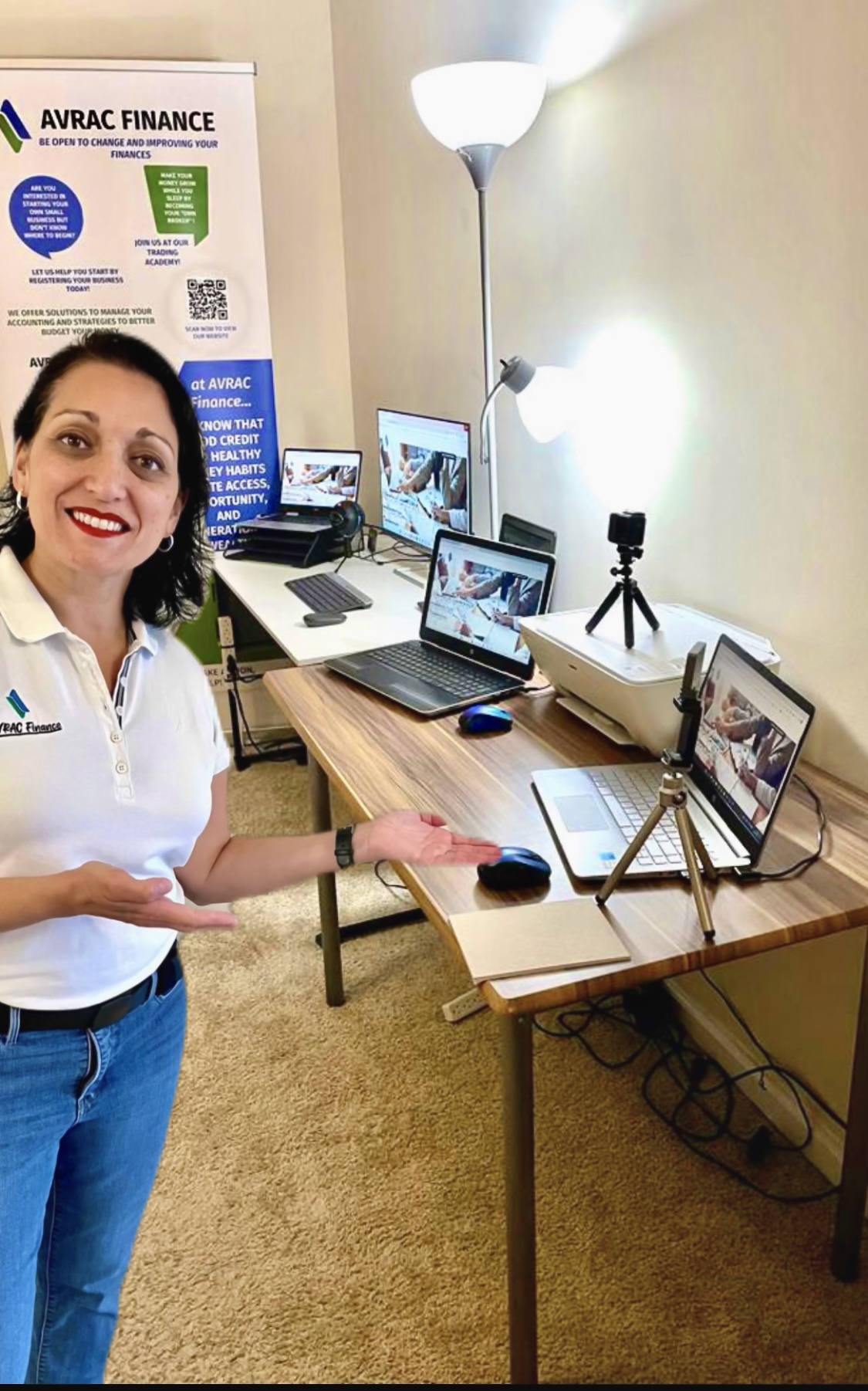 Woman smiling in office with laptops, tablets, and a tripod on a wooden table, and a floor lamp nearby.