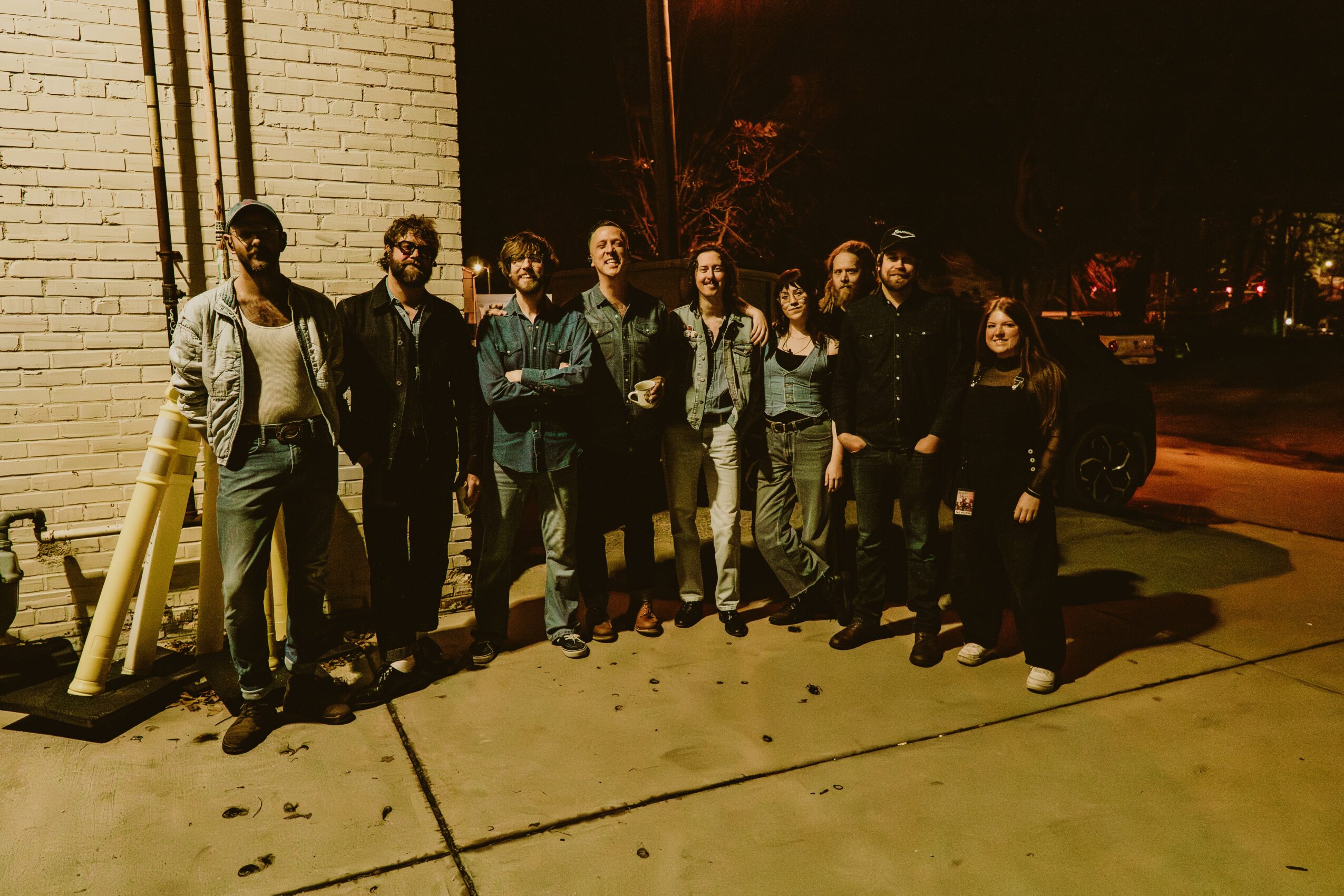 Group of nine people standing outside at night, illuminated by streetlights, with a brick wall and parked cars in background.