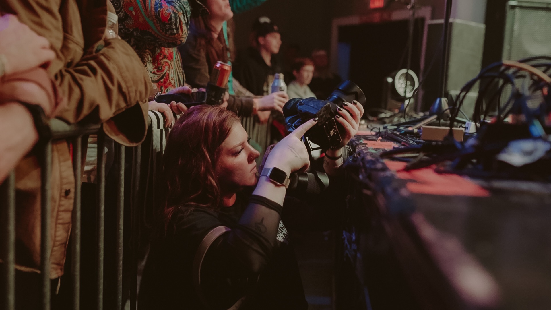 Group of people watching a woman taking photos at a concert or event, with equipment and screens in the background.