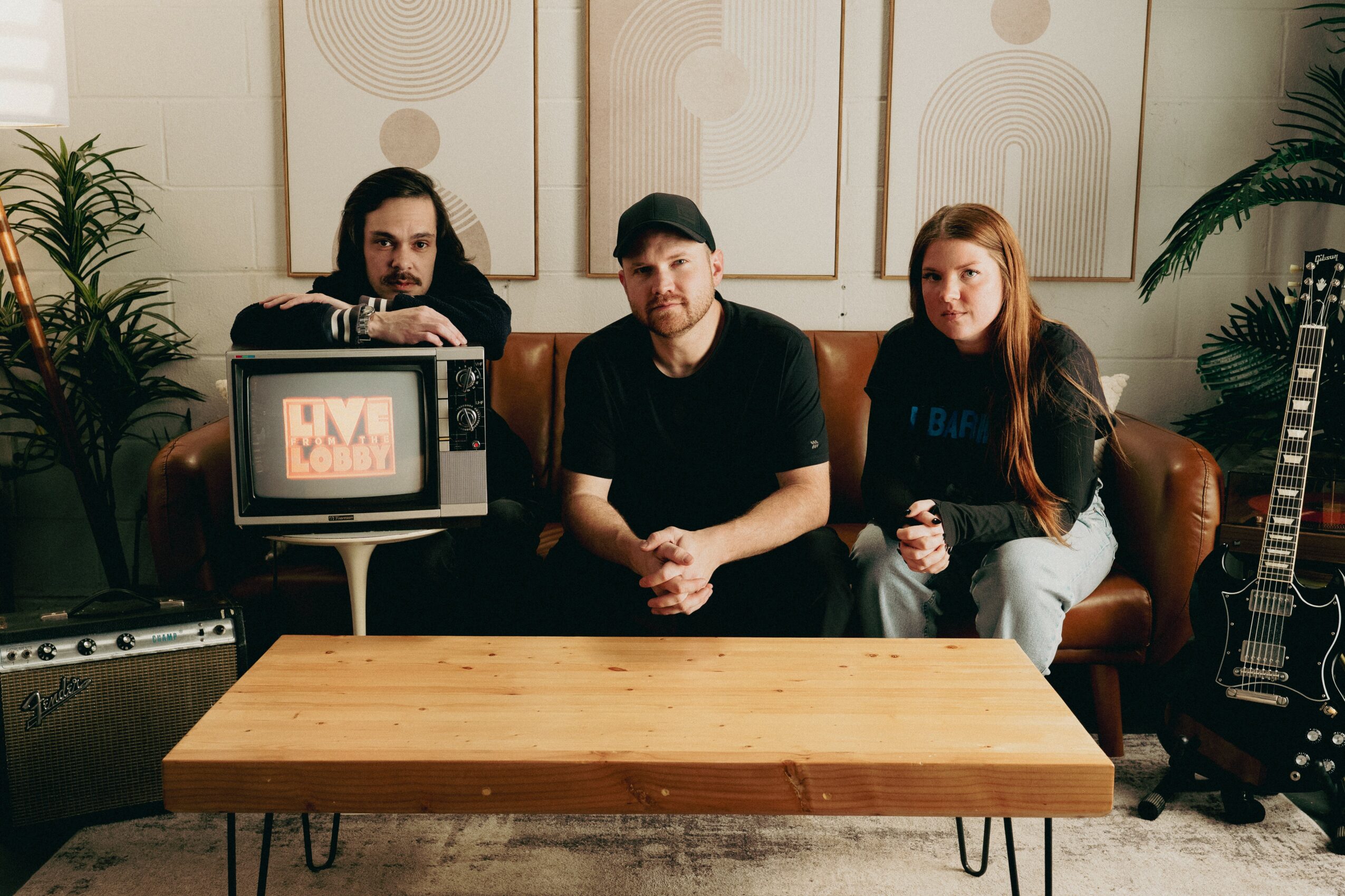 Three people sitting on a sofa in a room with plants and musical equipment, including a guitar and a monitor.