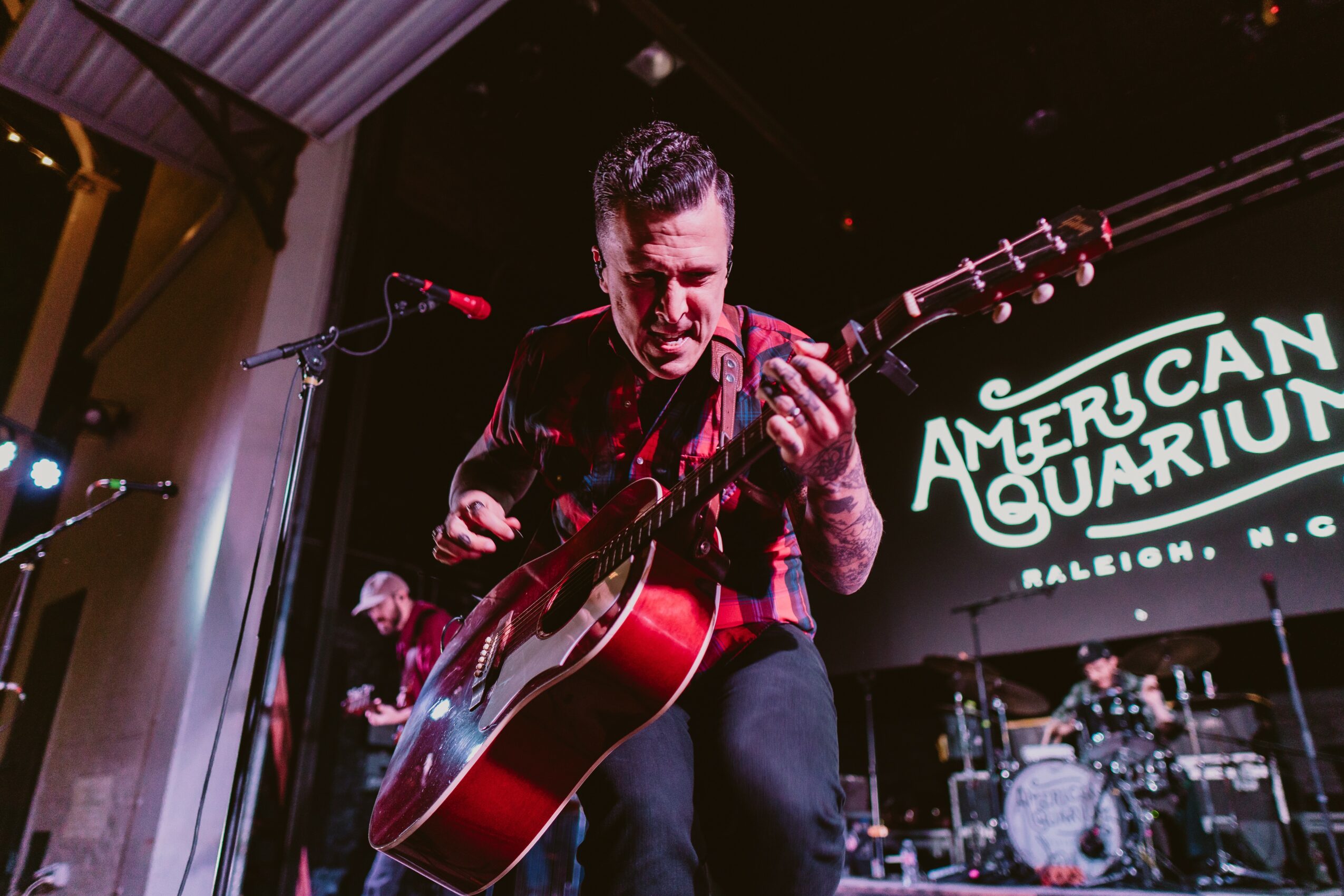 Musician playing guitar on stage with a band, illuminated by stage lights, with a sign reading 'American Aquarium' in the background.
