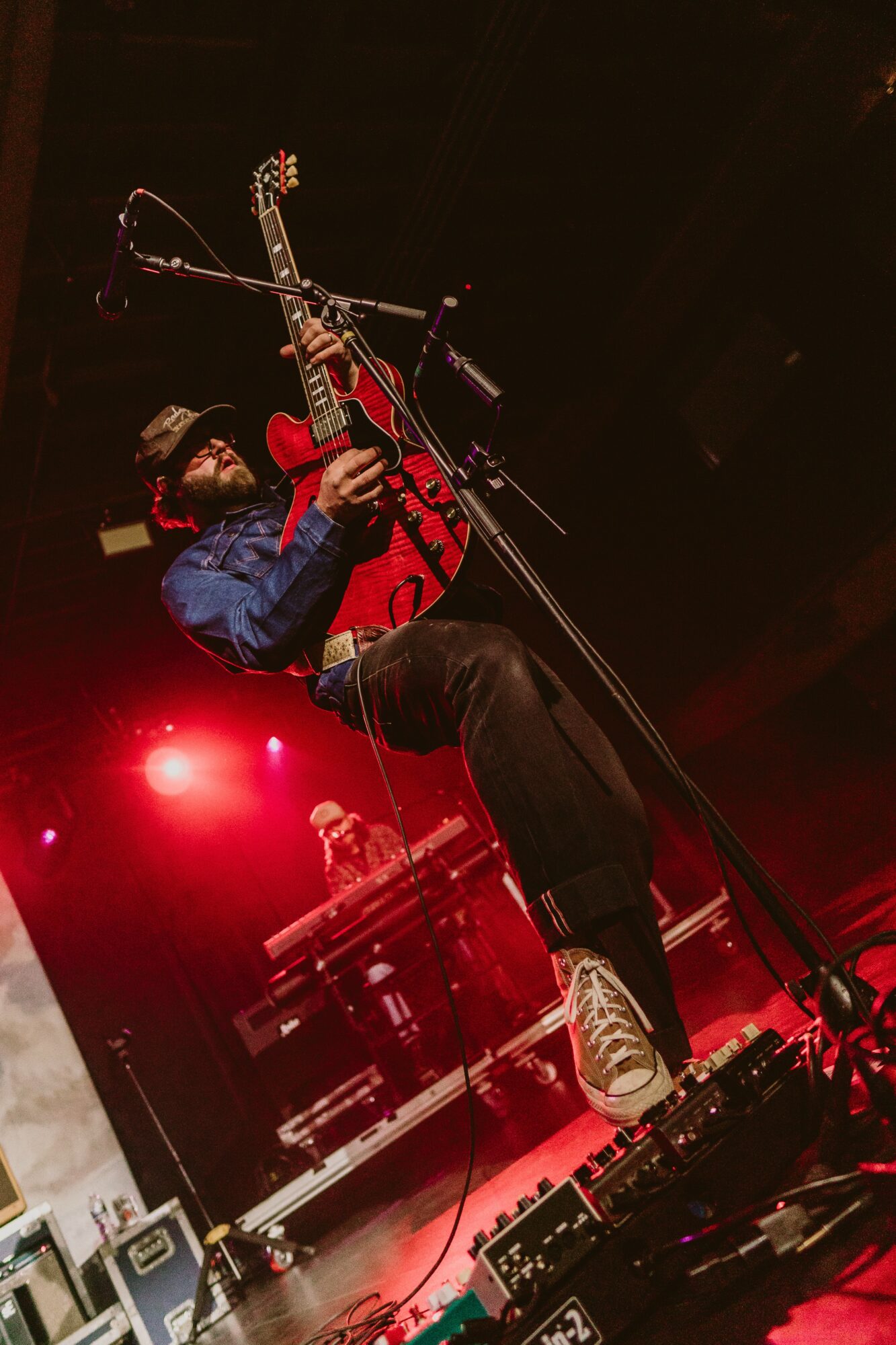 Musician playing guitar on stage with red lighting, wearing a cap, jacket, and sneakers, with equipment in the background.