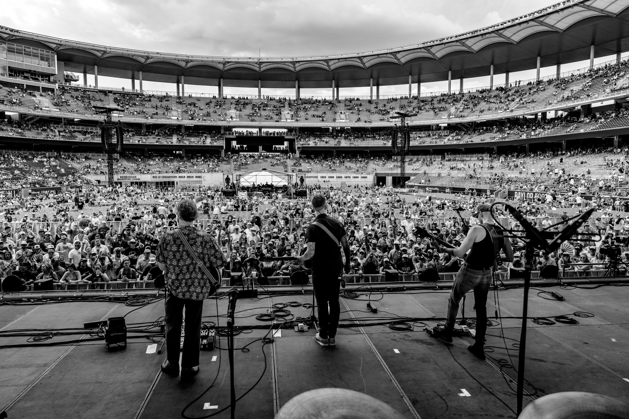 Band performing on stage in a large stadium filled with audience, black and white photo, stage equipment visible.