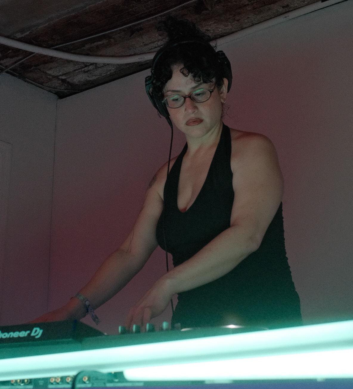 Woman with short dark hair and glasses, wearing a black top, DJing with equipment in a dimly lit room.