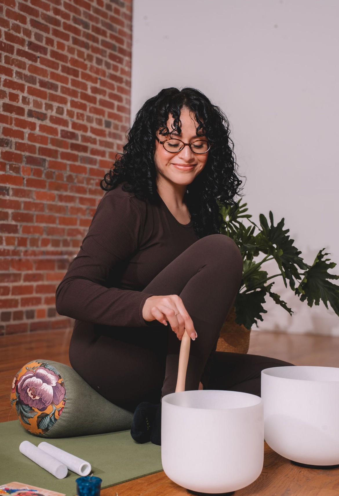 Woman with curly hair and glasses sits on a yoga mat, playing crystal singing bowls, with a brick wall and plant behind her.