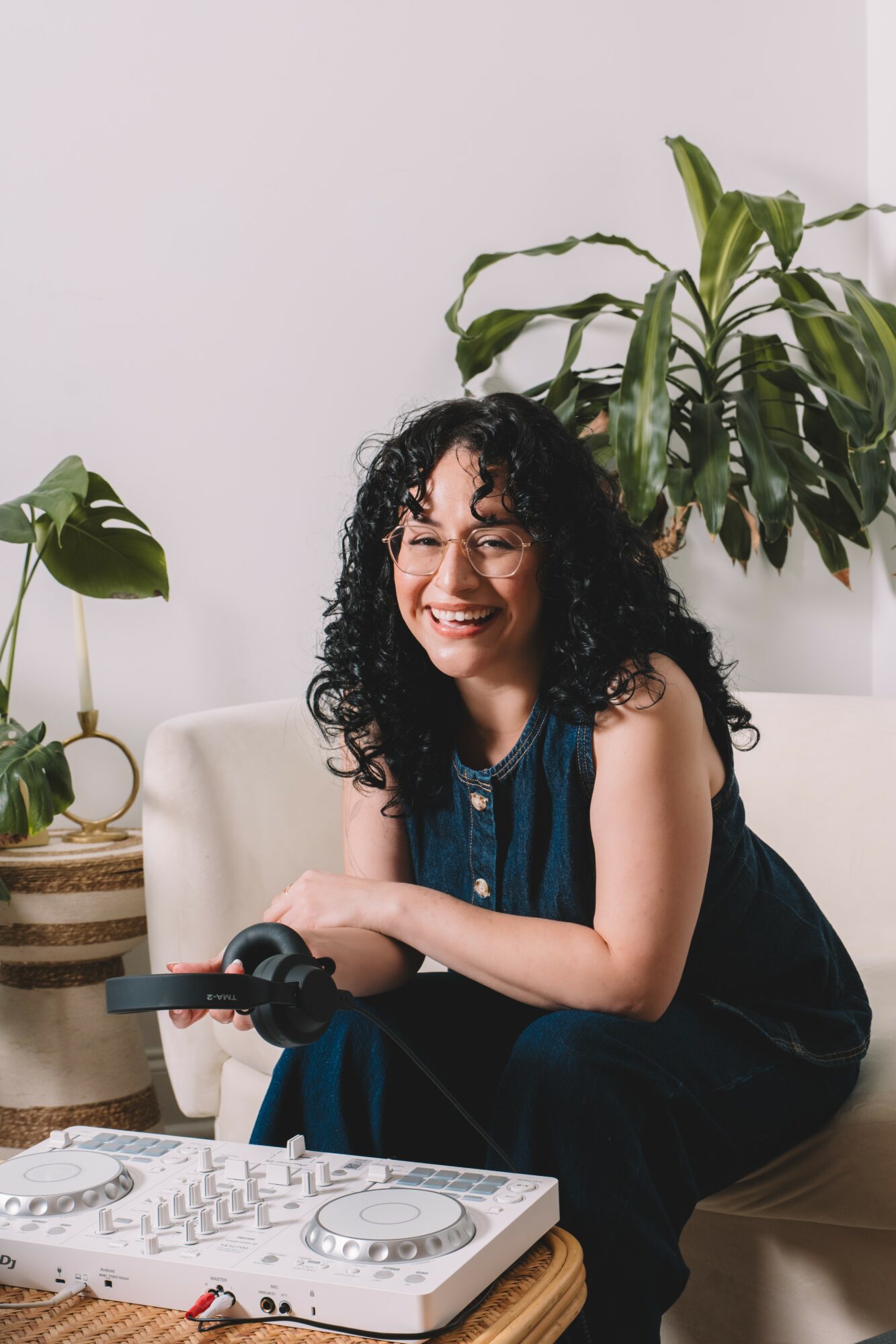 Woman with curly hair smiling, sitting on a beige sofa, surrounded by green plants in a bright room.