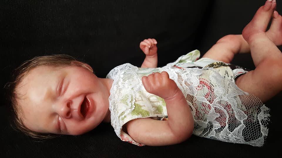 Baby lying on a dark surface, crying with eyes closed and fists clenched, wearing a lace outfit.