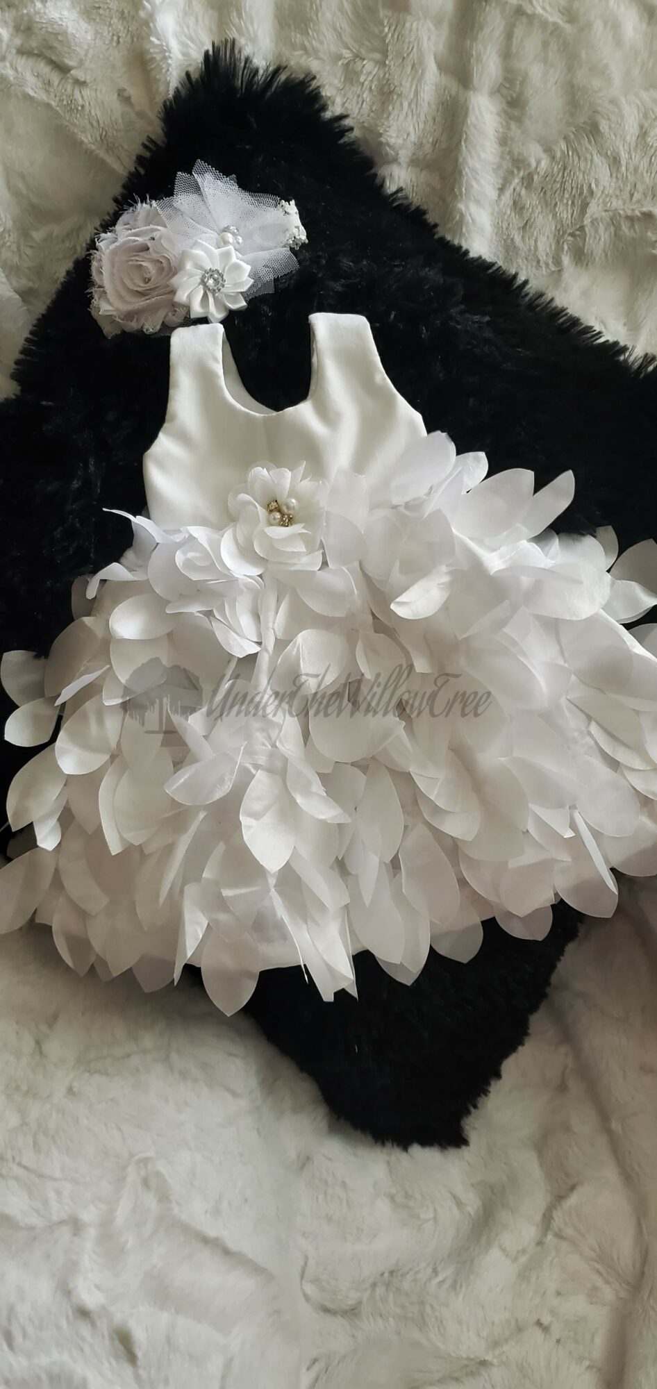 White floral dress with ruffled skirt on a black furry surface, with a small white flower accessory nearby.