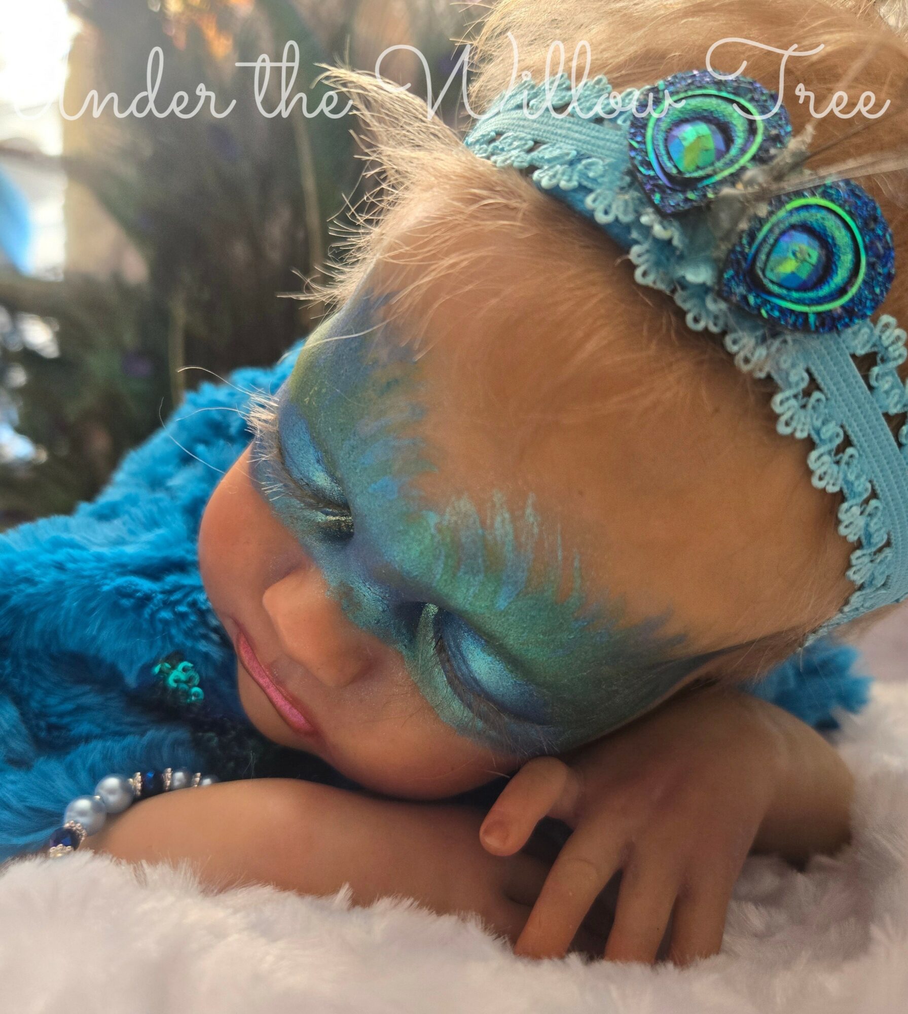 Child with peacock feather headband and face paint, resting on a soft surface outdoors, with text 'under the Willow Tree' at top.