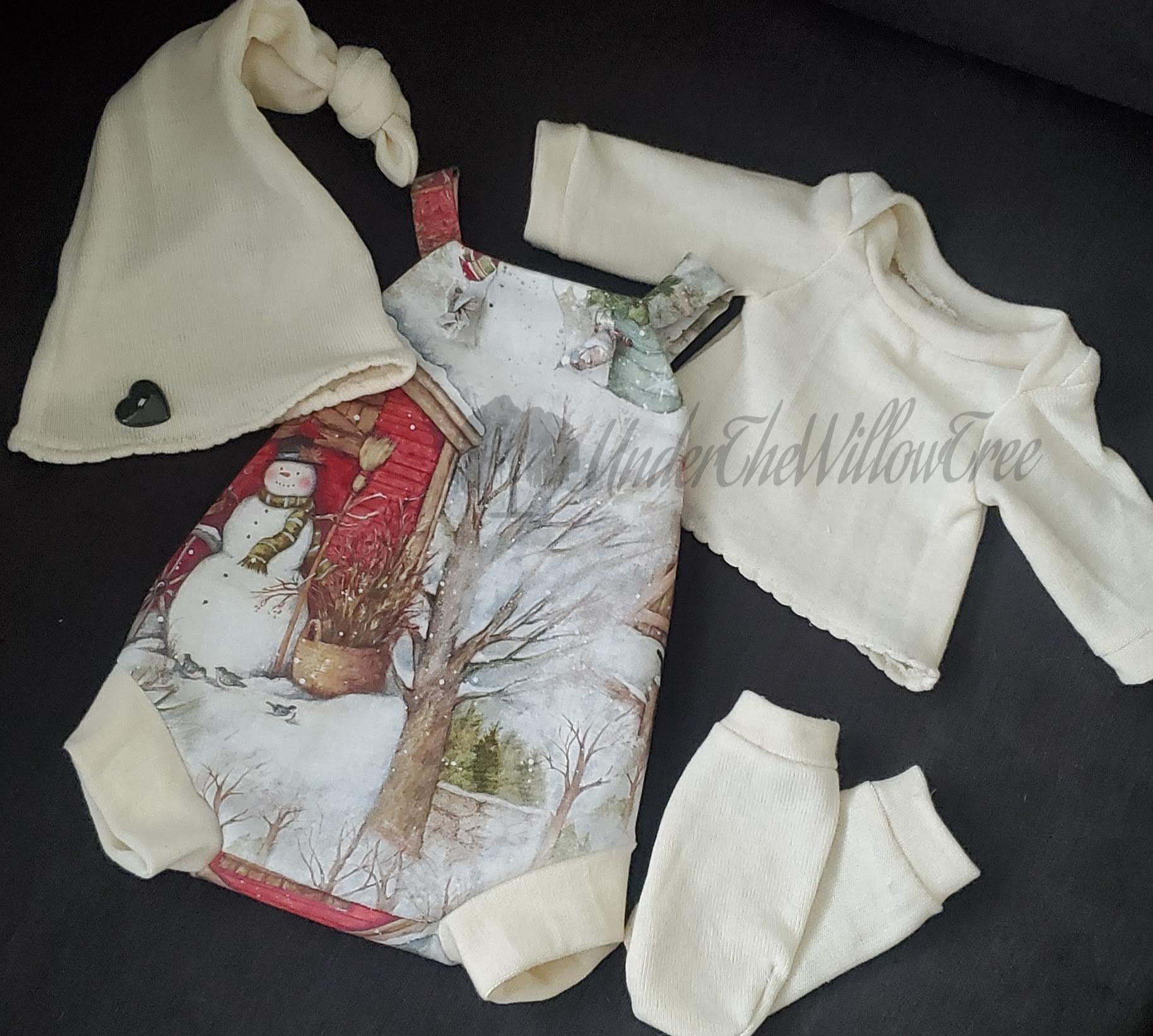 Baby onesie with winter scene, cream hat with knot, and matching mittens on black background.