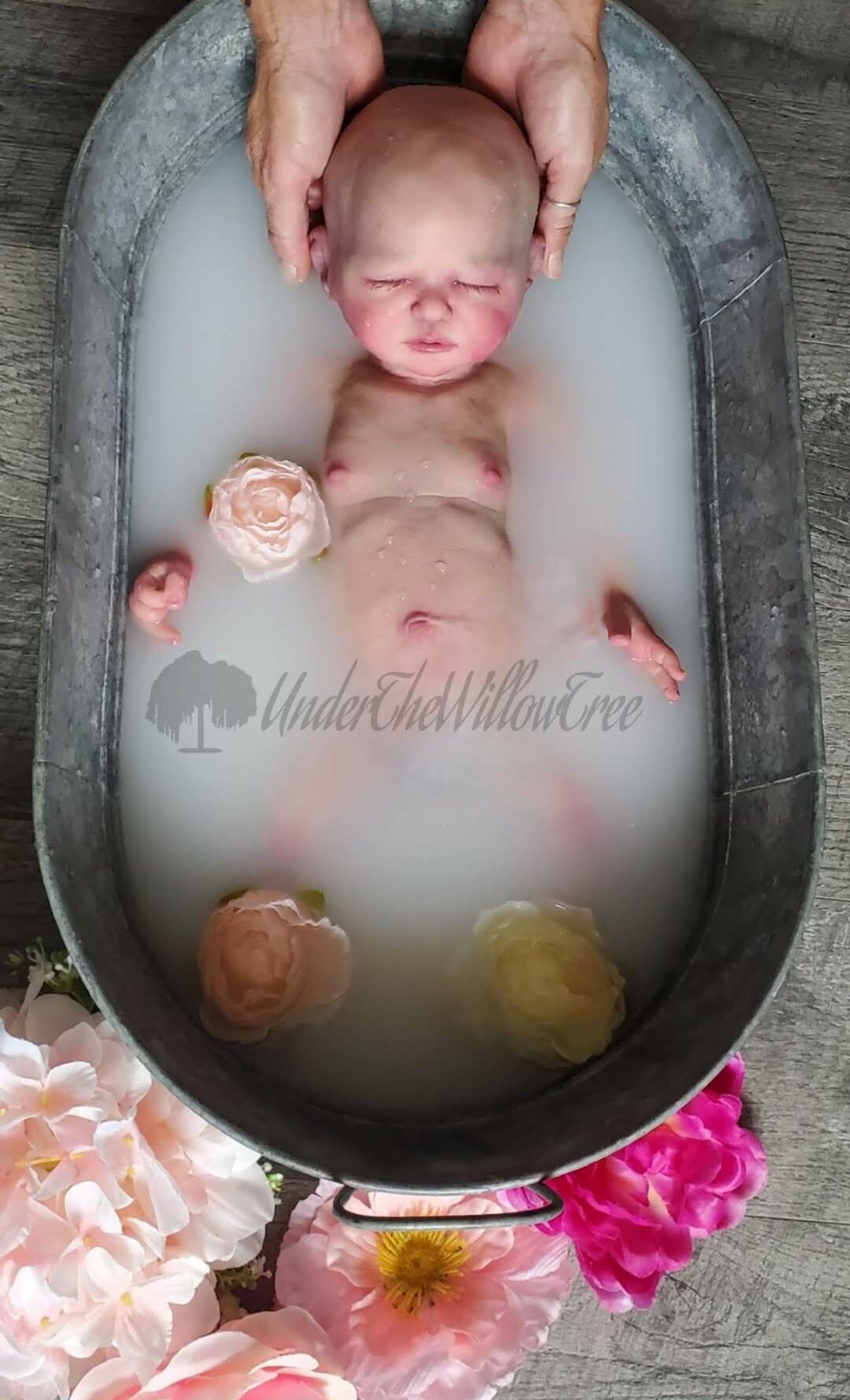 Baby receiving a milk bath in a black tub, surrounded by flowers, with hands gently supporting the head.