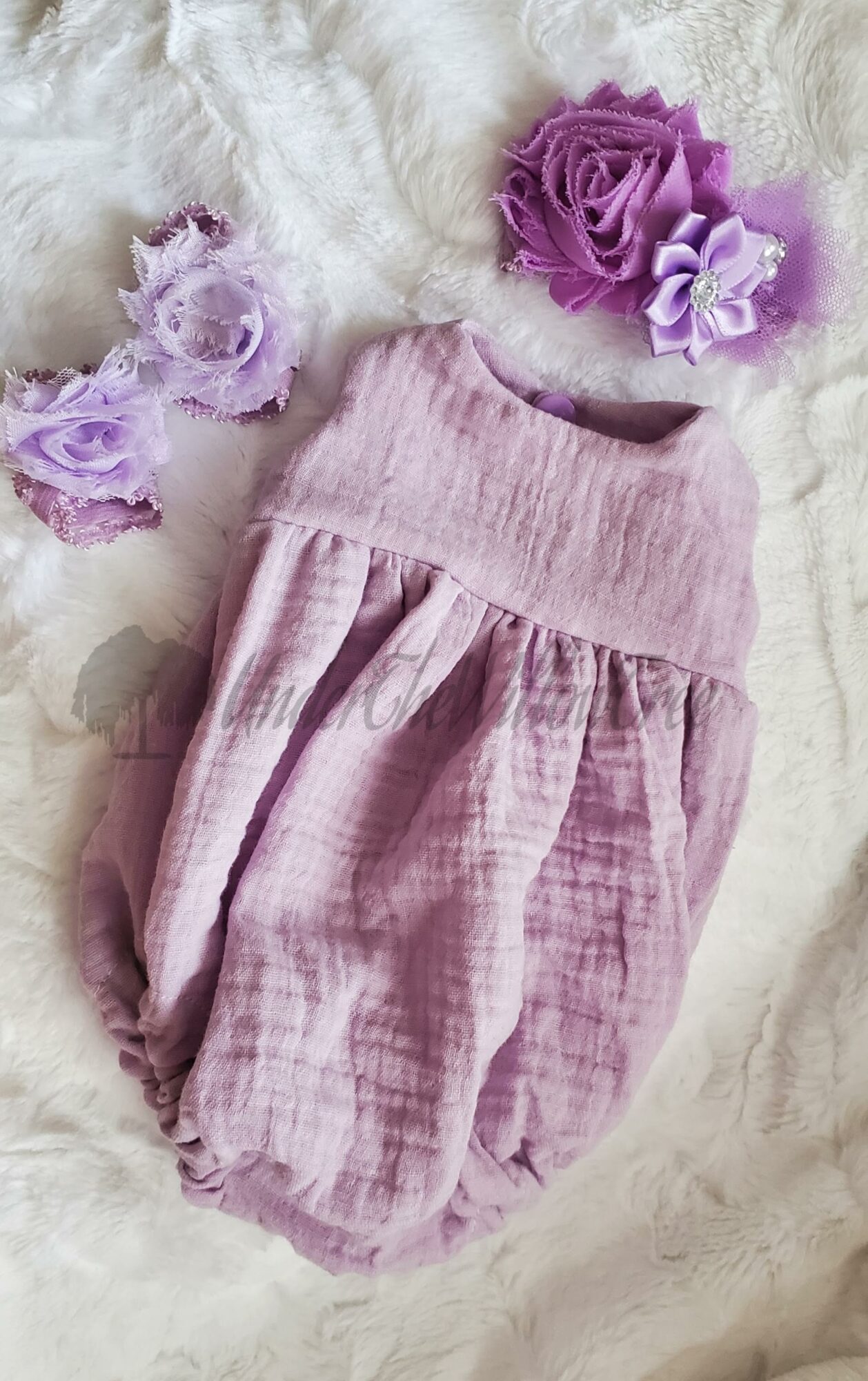 Pink baby romper with gathered waist, surrounded by purple flowers on a soft surface.