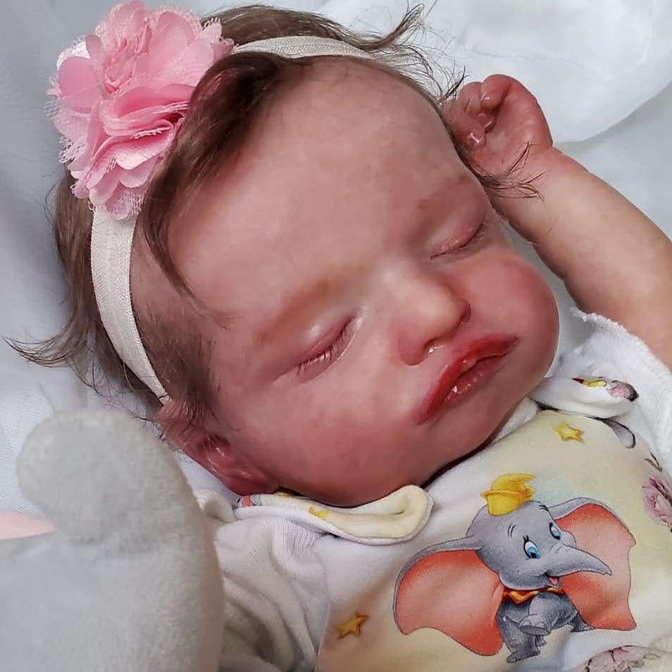 Baby with a pink flower headband sleeping, resting head on a white surface, wearing a shirt with cartoon elephant, 20 words