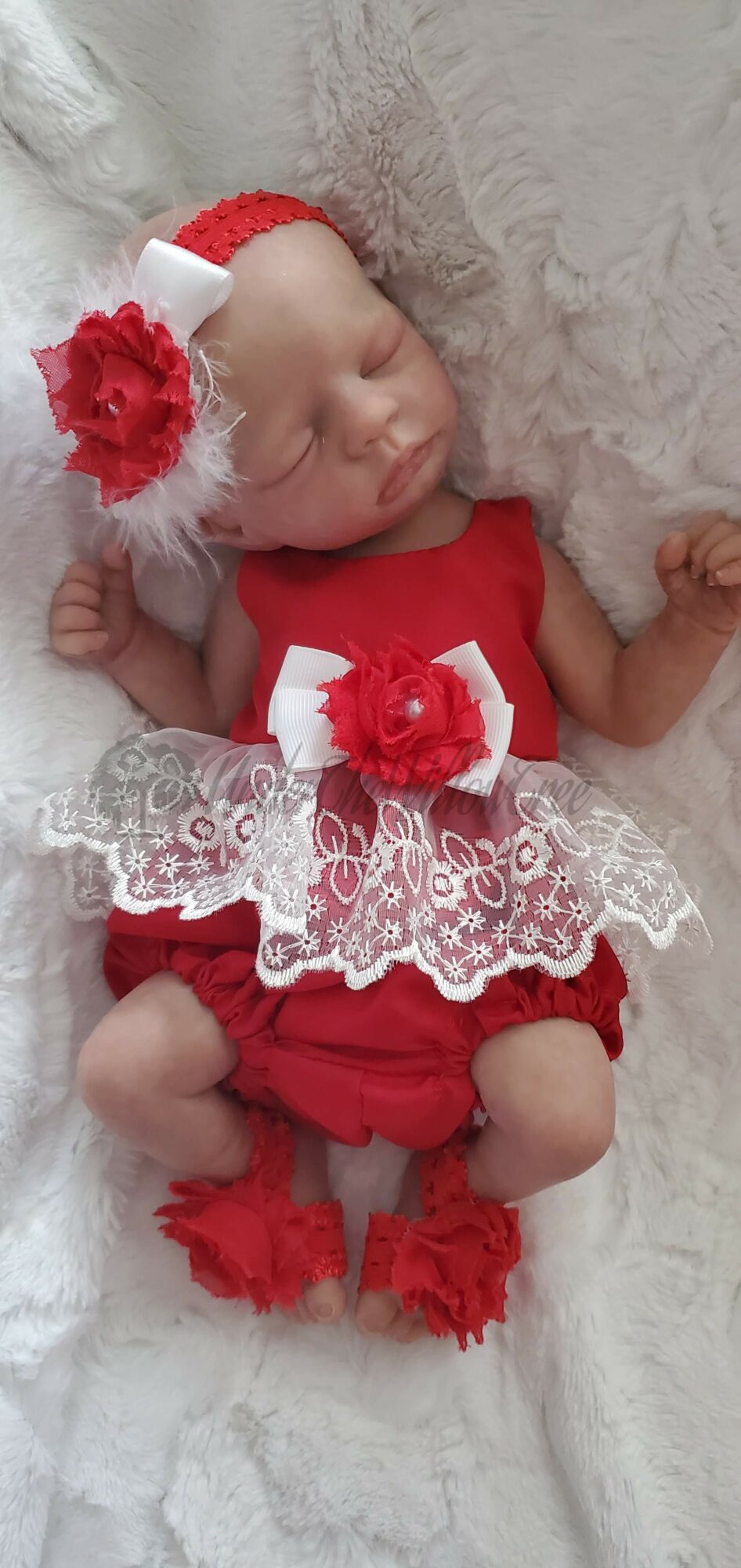 Baby dressed in red with lace and flower accessories, sleeping on a soft surface.