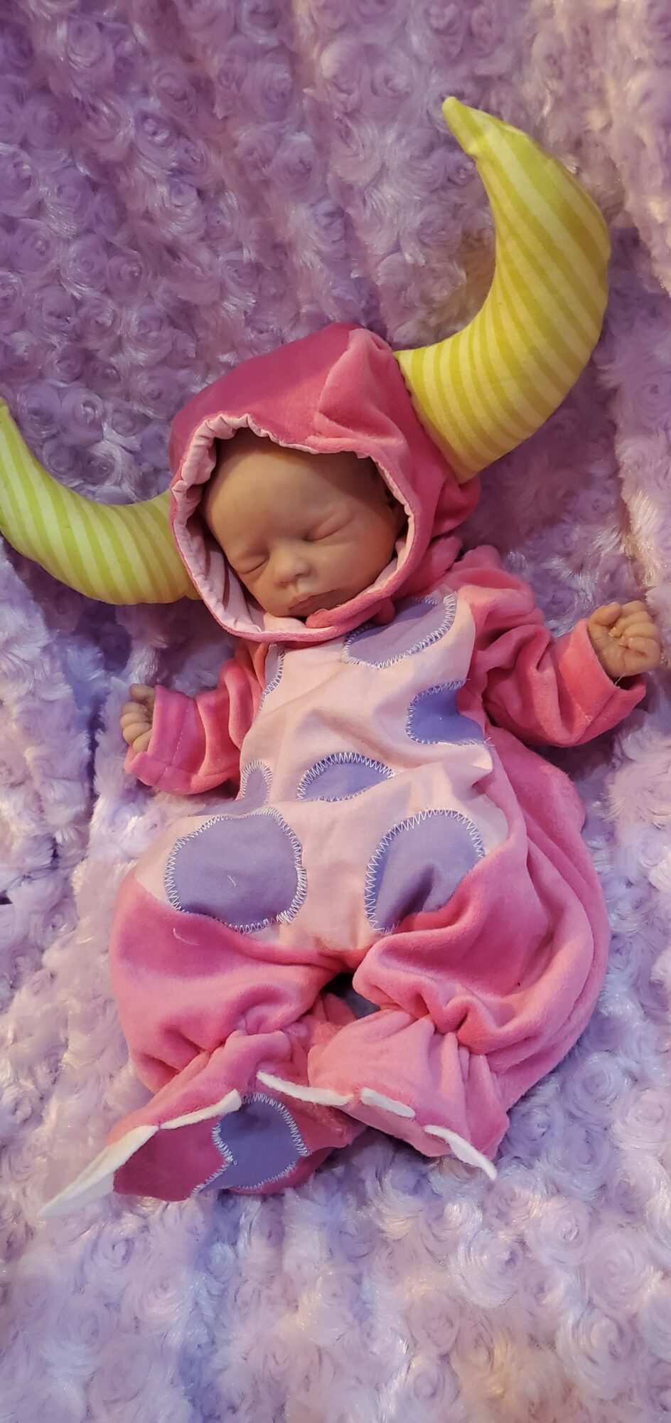 Baby in pink costume with yellow horns, lying on purple fluffy surface, eyes closed, wrapped in pink fabric.