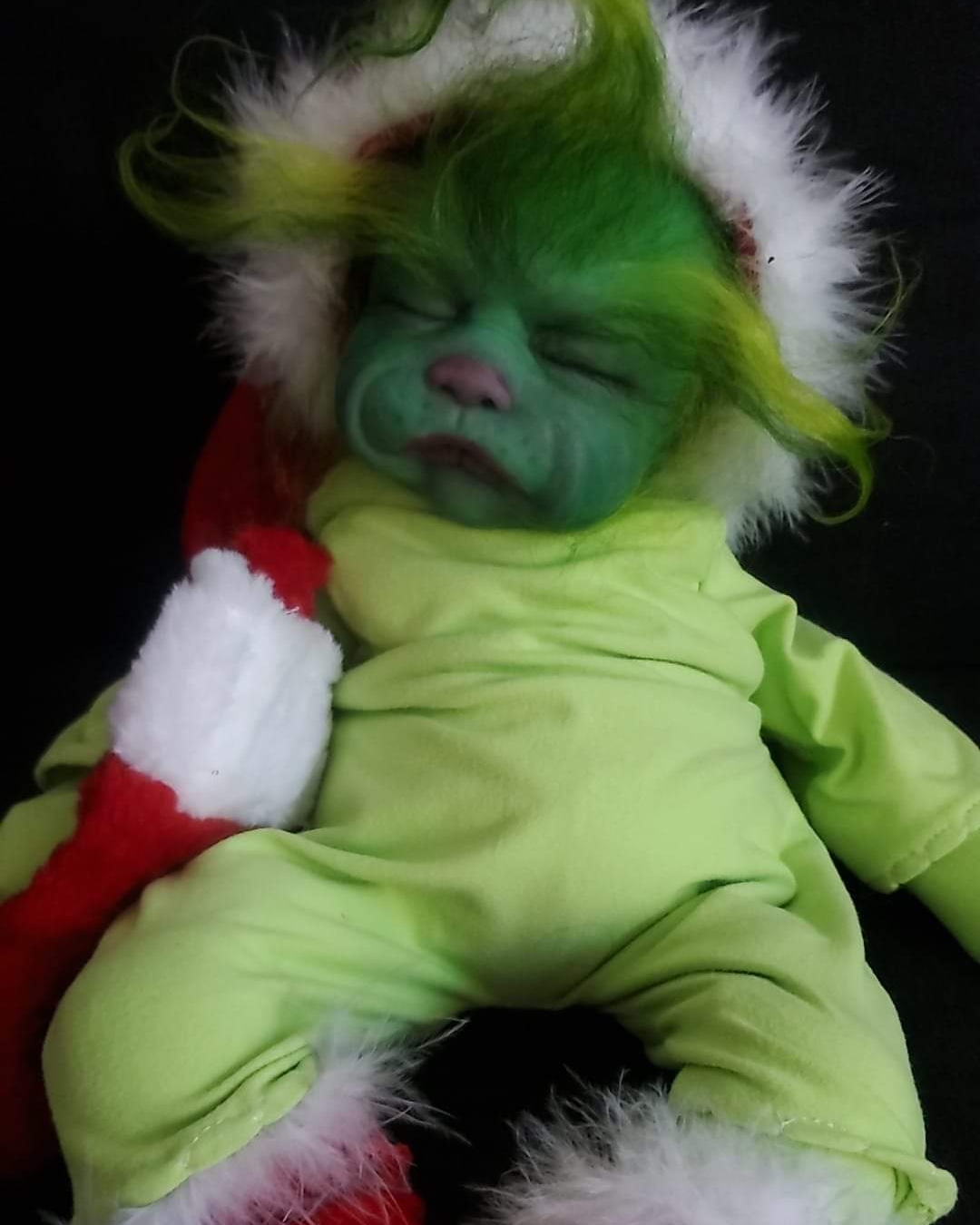 Child dressed as a green creature with face paint, wearing a Santa hat and a yellow-green outfit, lying down with eyes closed.