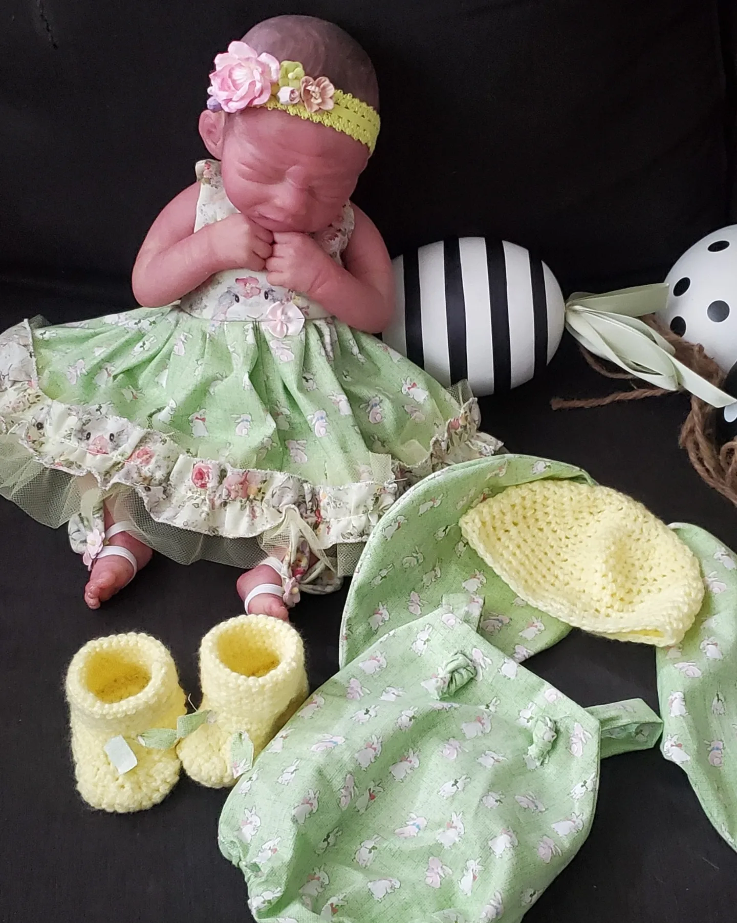 Baby girl wearing a floral headband and green dress, sitting with yellow booties and decorative objects nearby.