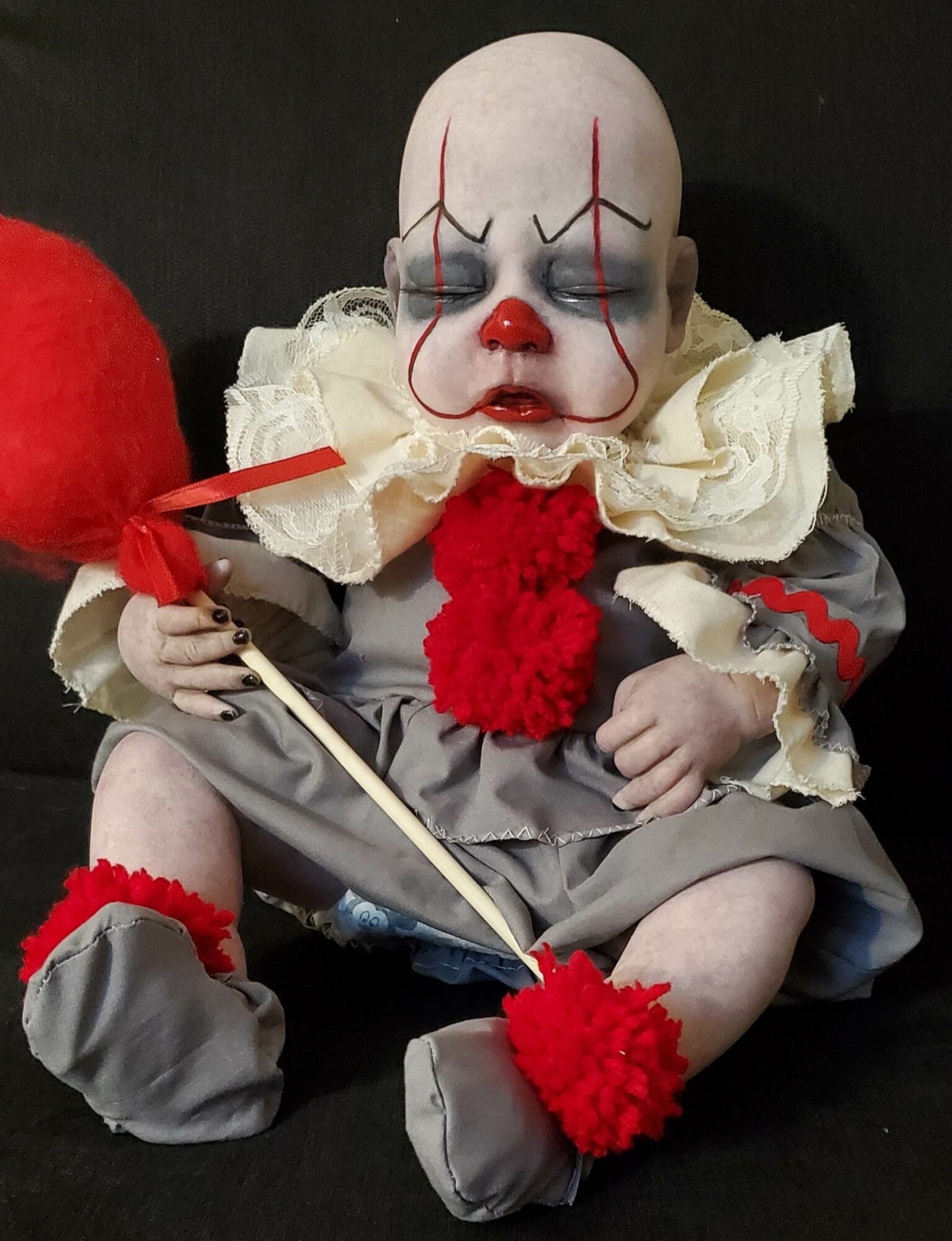 Creepy clown doll with painted face, ruffled collar, holding a red balloon, dressed in gray and red, sitting against black background.
