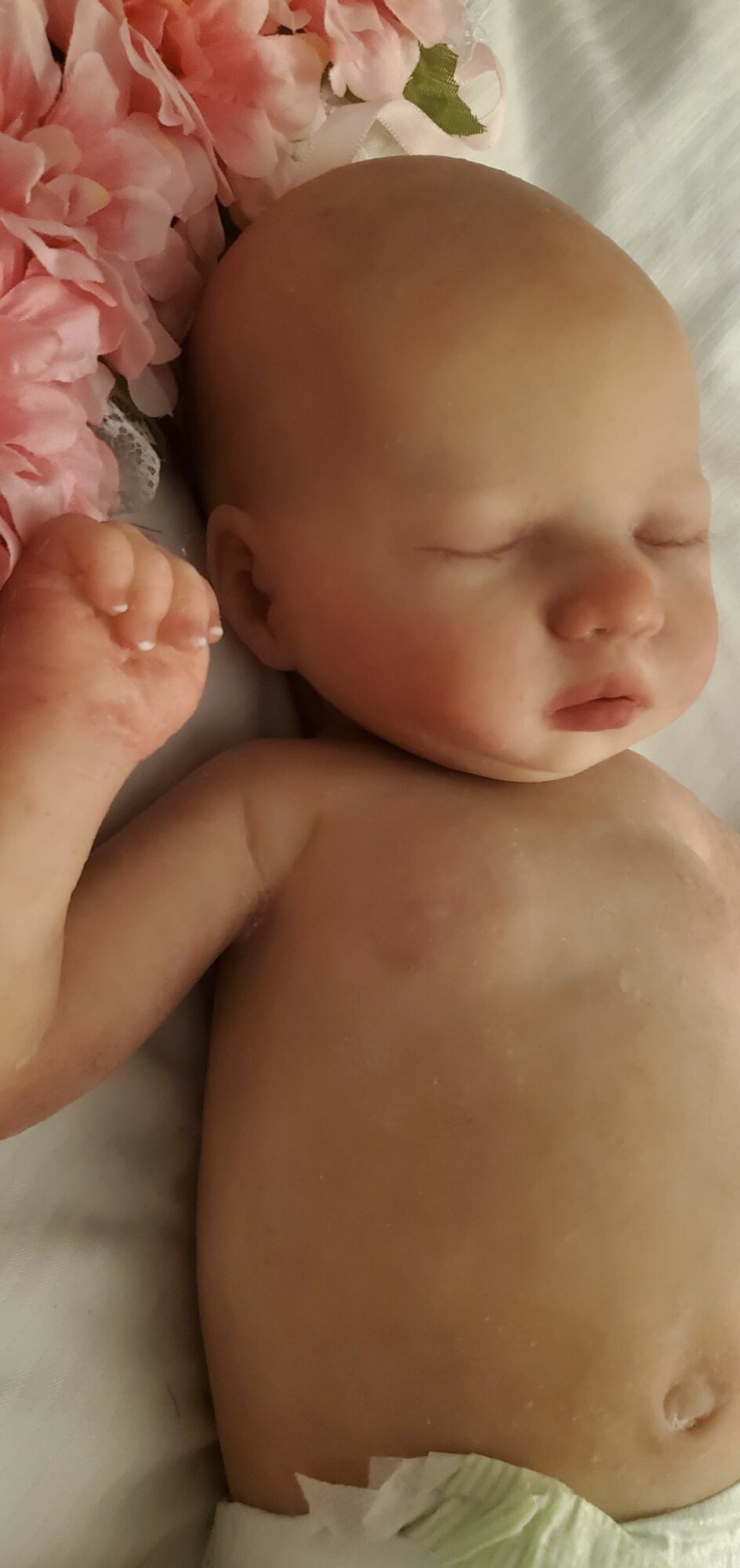 A sleeping baby lying on a soft surface with pink flowers nearby, eyes closed, and arms relaxed.